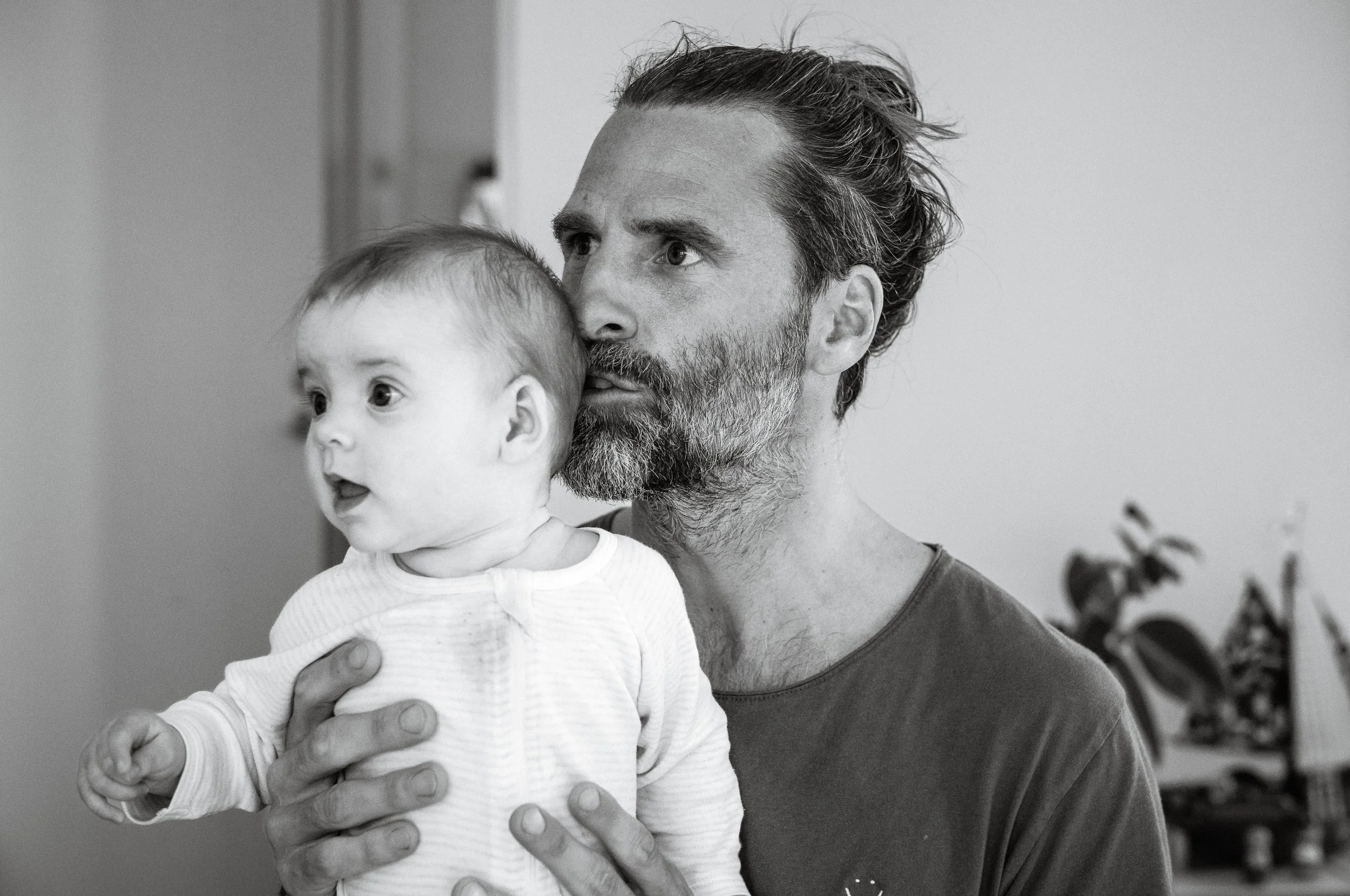 A man with a beard holding a baby with light hair, both looking surprised or curious in an indoor setting.