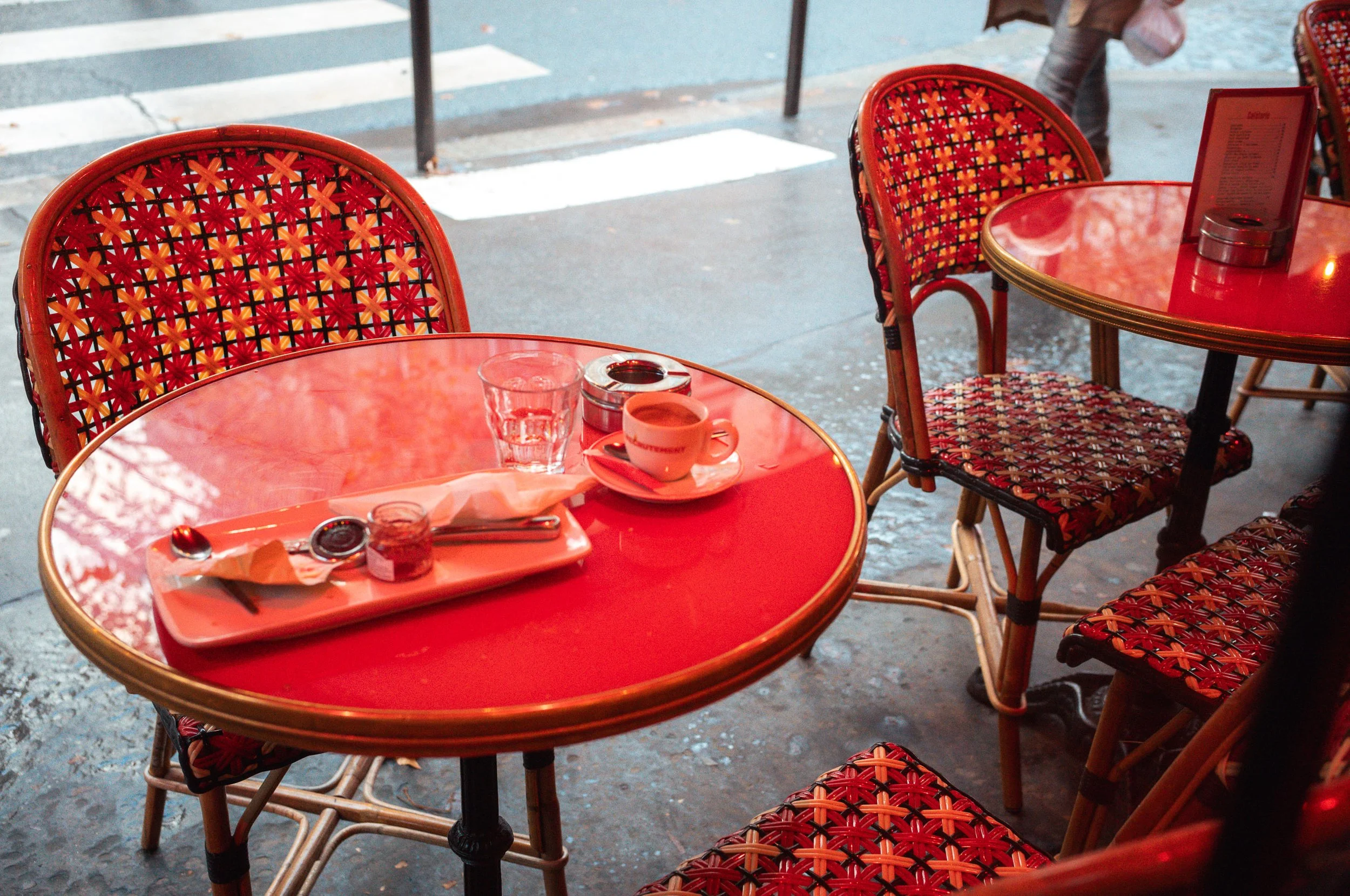Outdoor cafe table with a red surface, set with a small cup of coffee, a glass of water, a small jar, and utensils on a tray, surrounded by matching patterned chairs, on a city sidewalk.