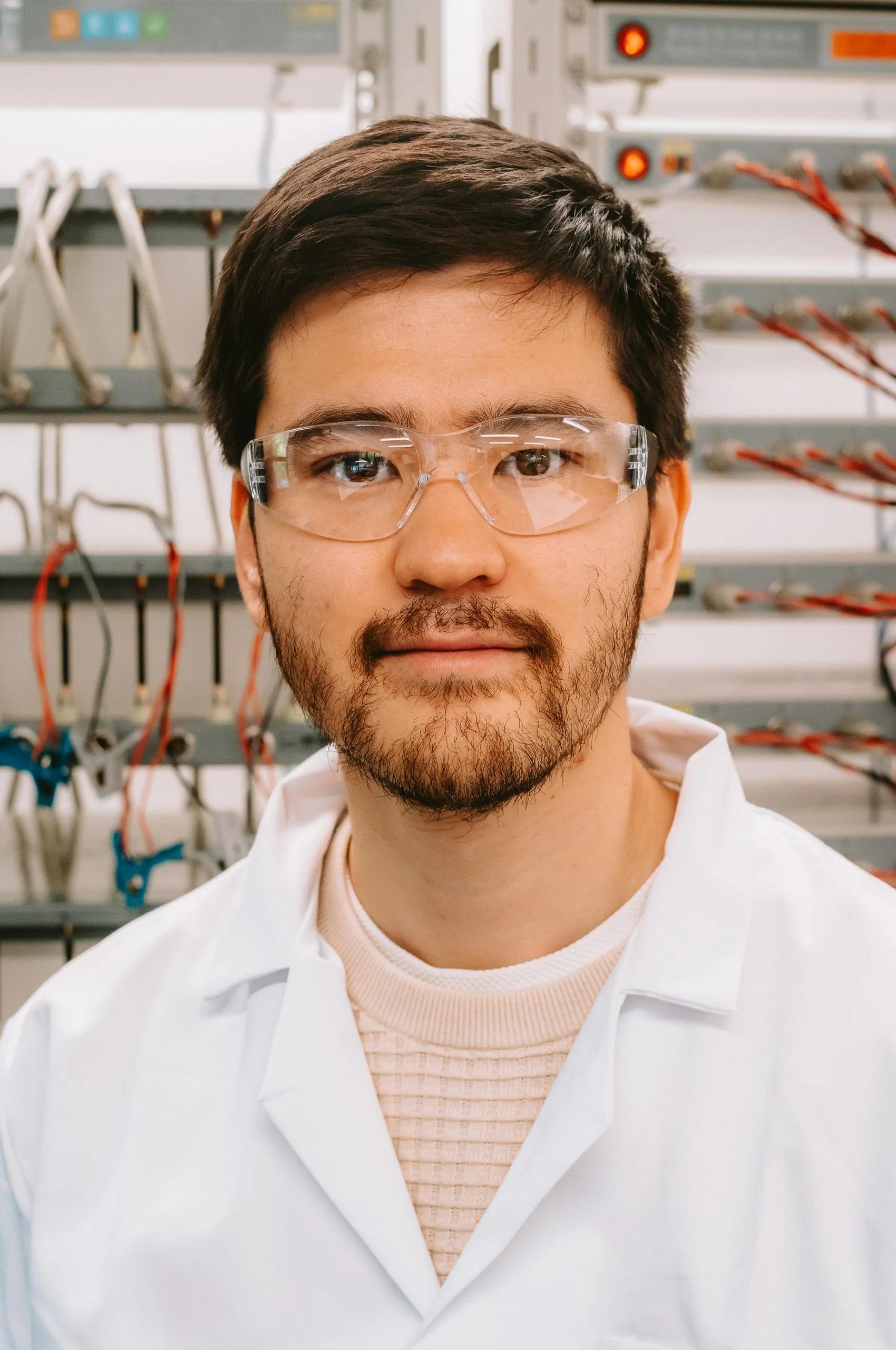 A scientist in safety glasses and a lab coat standing in a laboratory with scientific equipment and wires in the background.