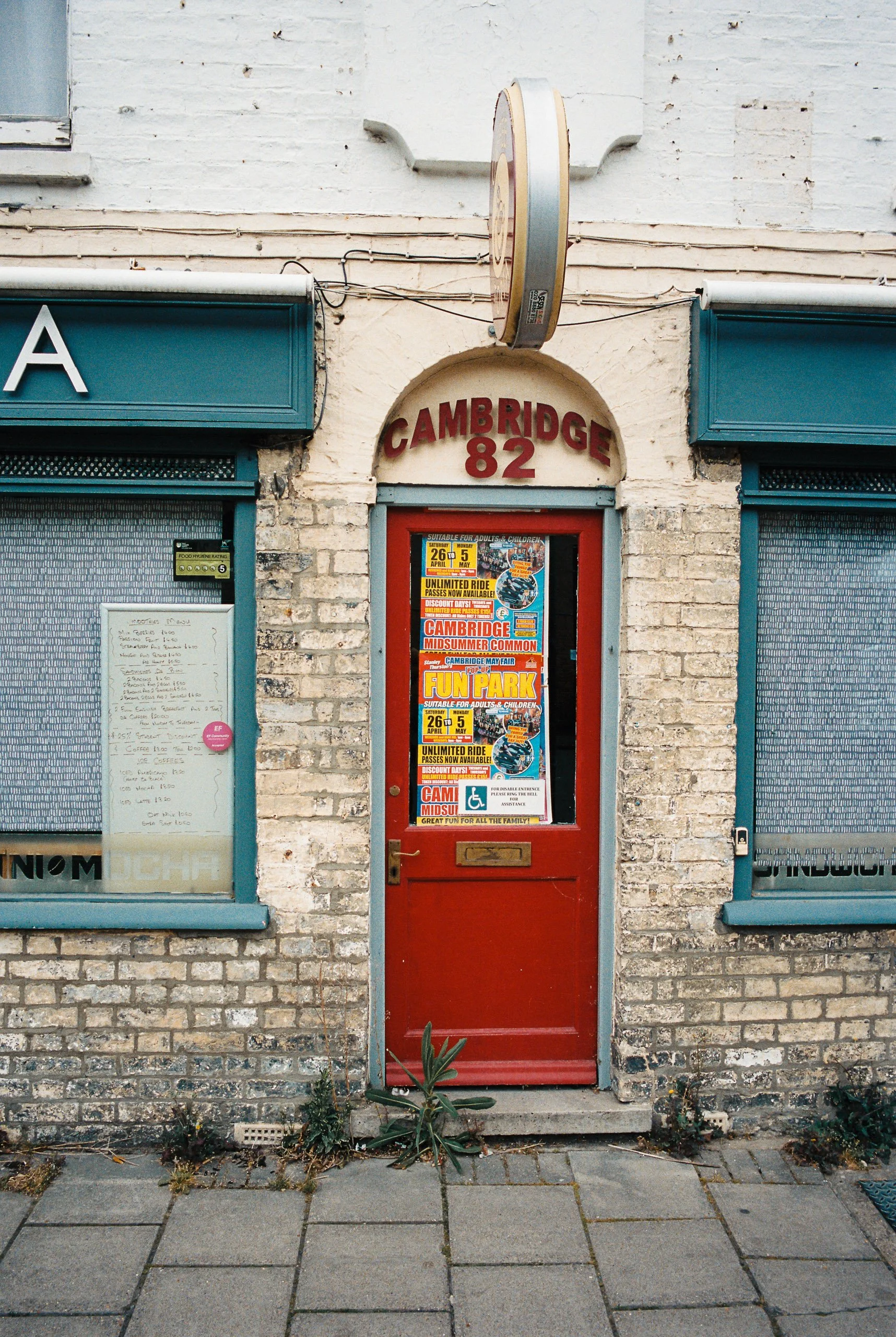 Red door with posters advertising Cambridge fun park and school events, above a brick building with blue window frames, located at 82 Cambridge Street.