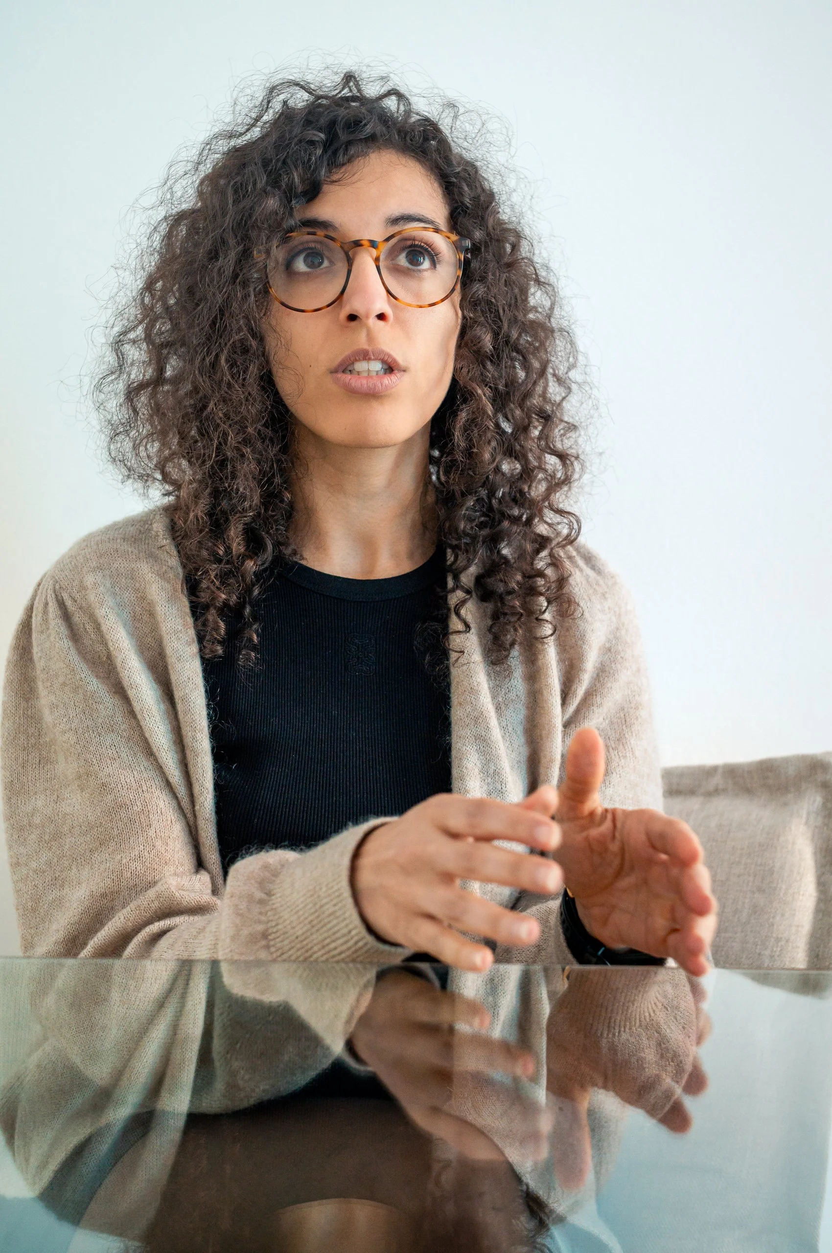 A woman with curly brown hair, wearing glasses, a black shirt, and a beige cardigan, sitting at a table and gesturing with her hands.