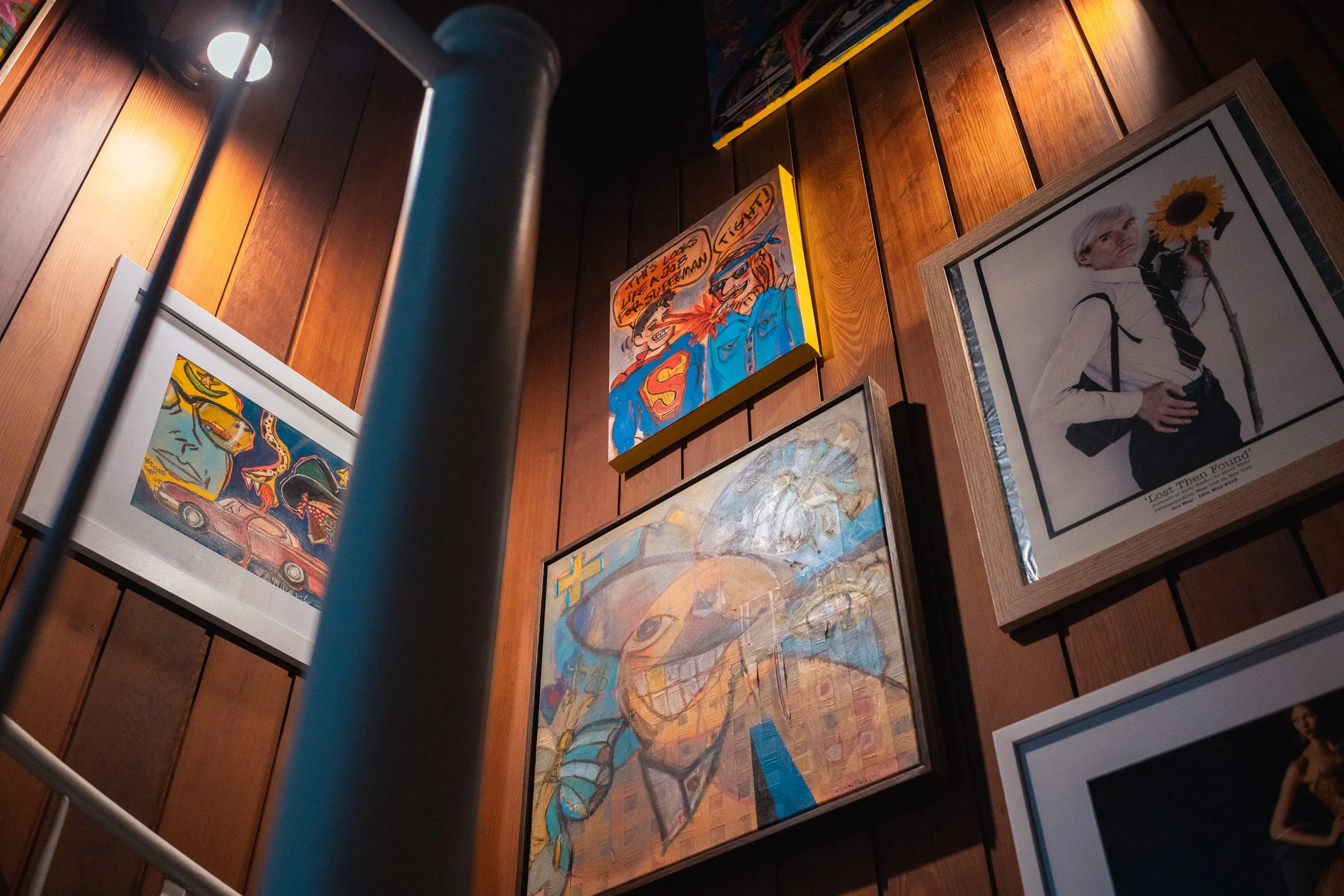 Art gallery wall with colorful paintings, including a comic-style Superman with police characters, a painted woman with an umbrella, and a woman with a sunflower, against wood-paneled wall.