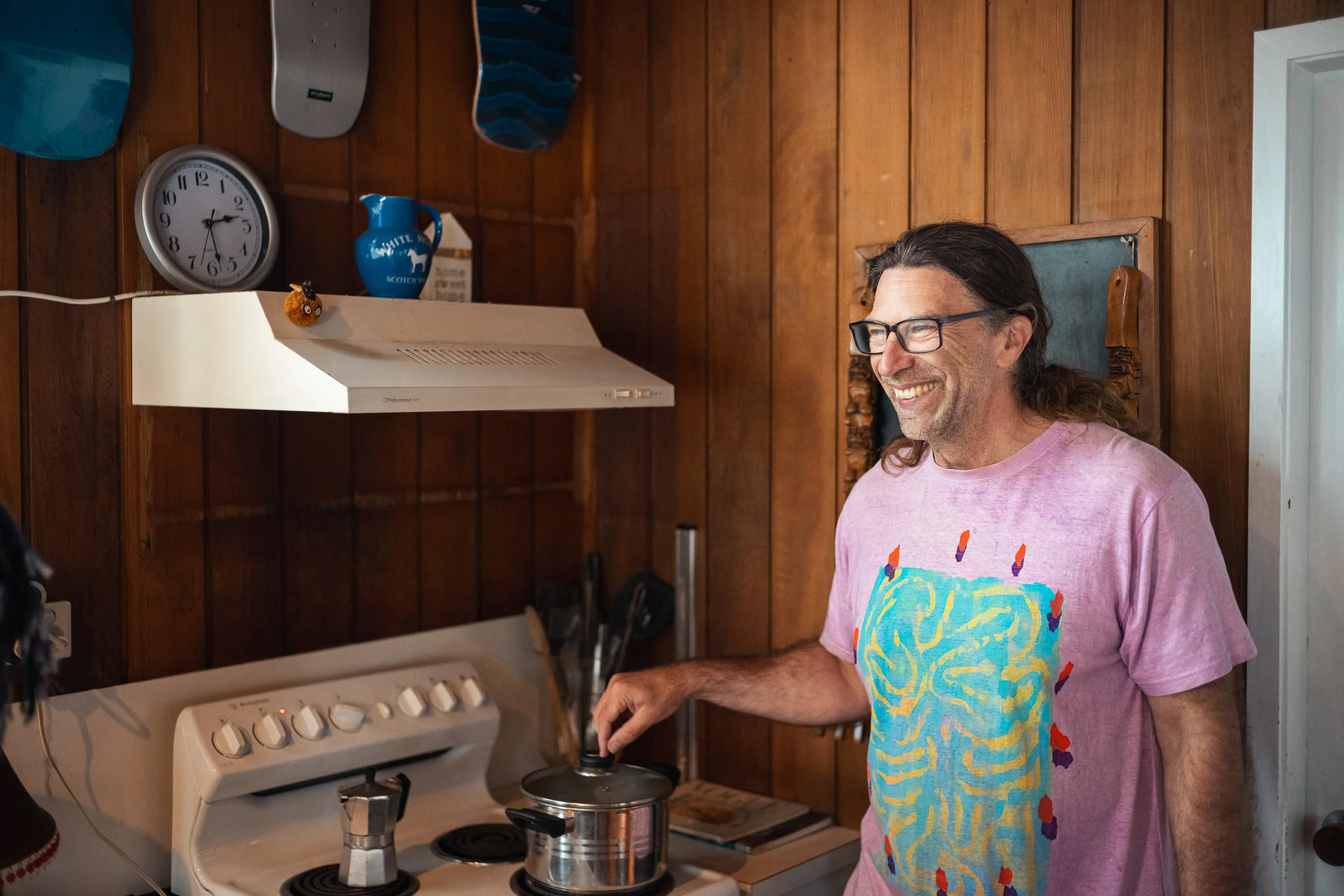 A man with long hair and glasses wearing a pink T-shirt with a colorful design, standing in a kitchen and smiling while stirring a pot on a stove.