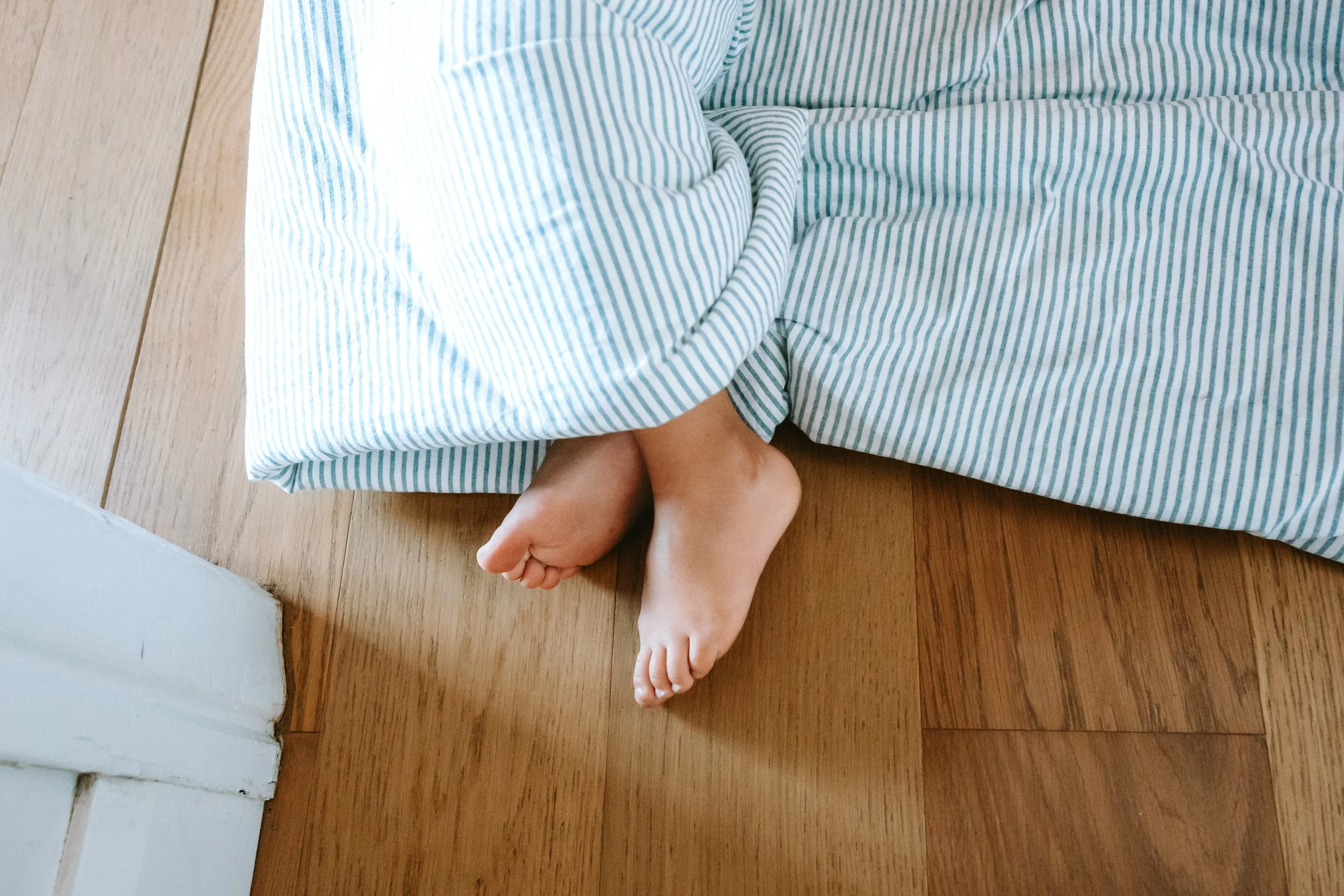 Close-up of a child's toes peeking out from under a horizontally striped blue and white blanket on a wooden floor.