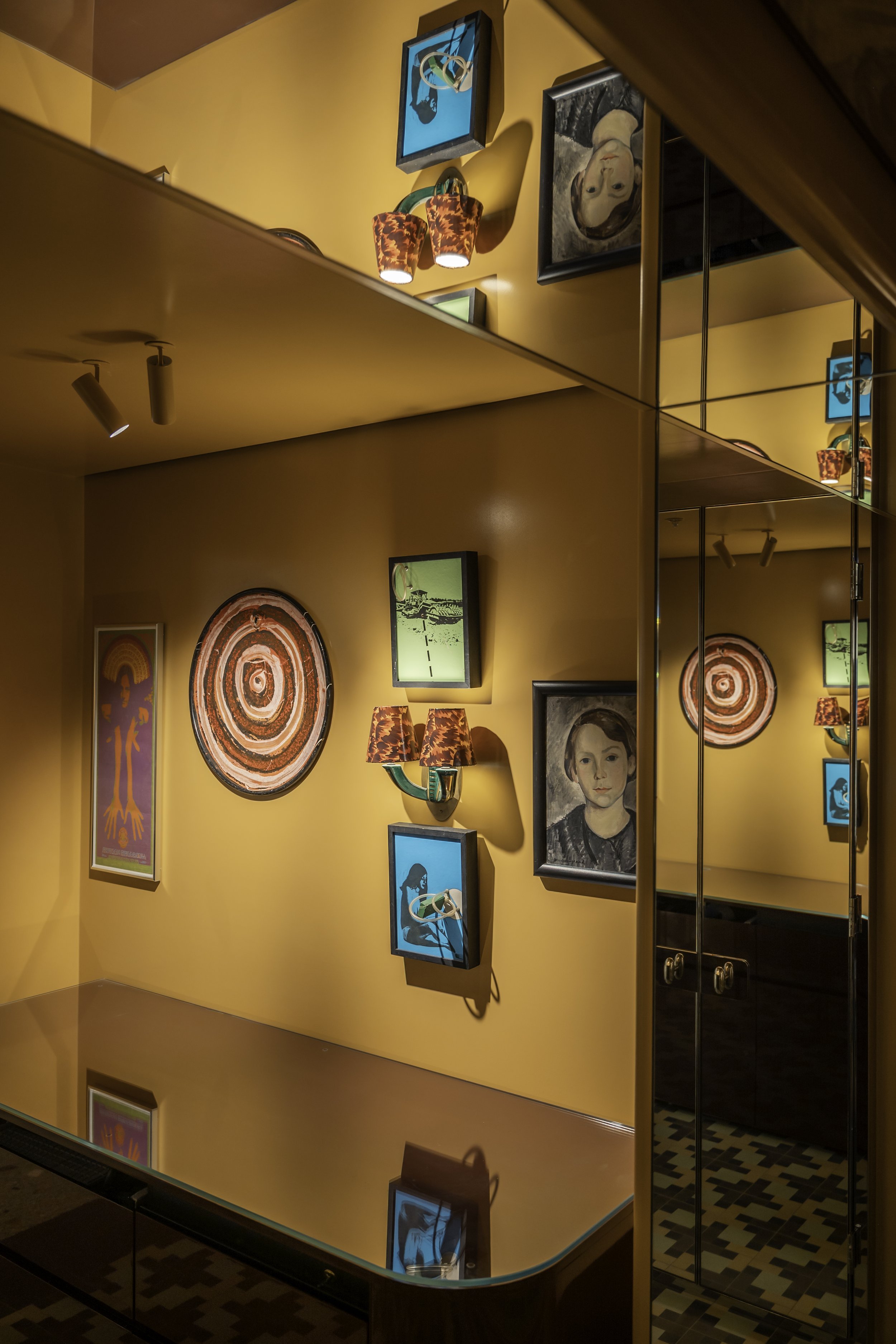 A reflection of a colorful art gallery wall with various framed paintings and decorative wall lamps, visible in a mirror.