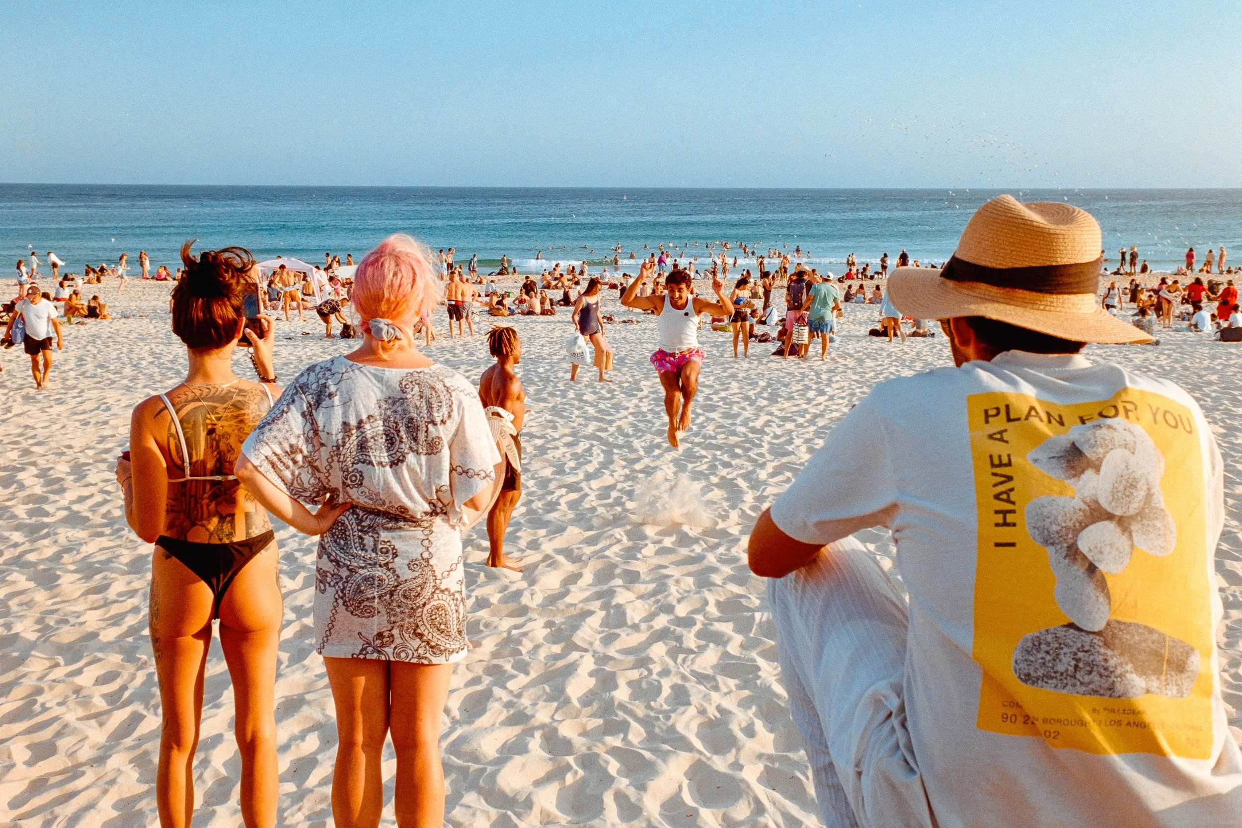 People on a crowded sandy beach, some watching a man jump mid-air, others taking photos, with ocean waves in the background and a clear sky.