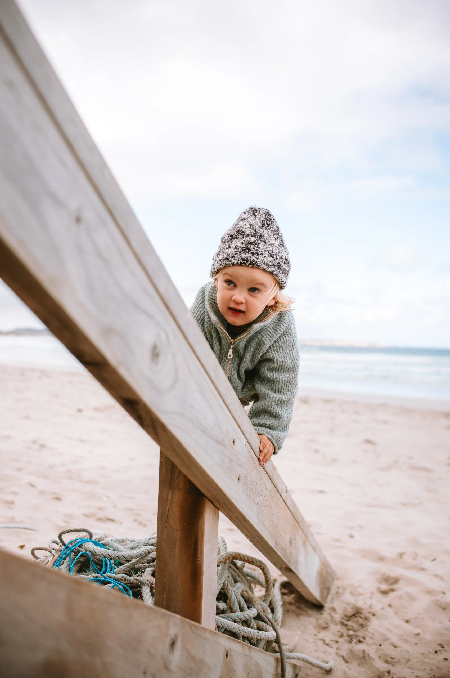 A young child climbing over a wooden barrier on a beach with sand and ocean in the background, wearing a knitted hat and gray sweater.