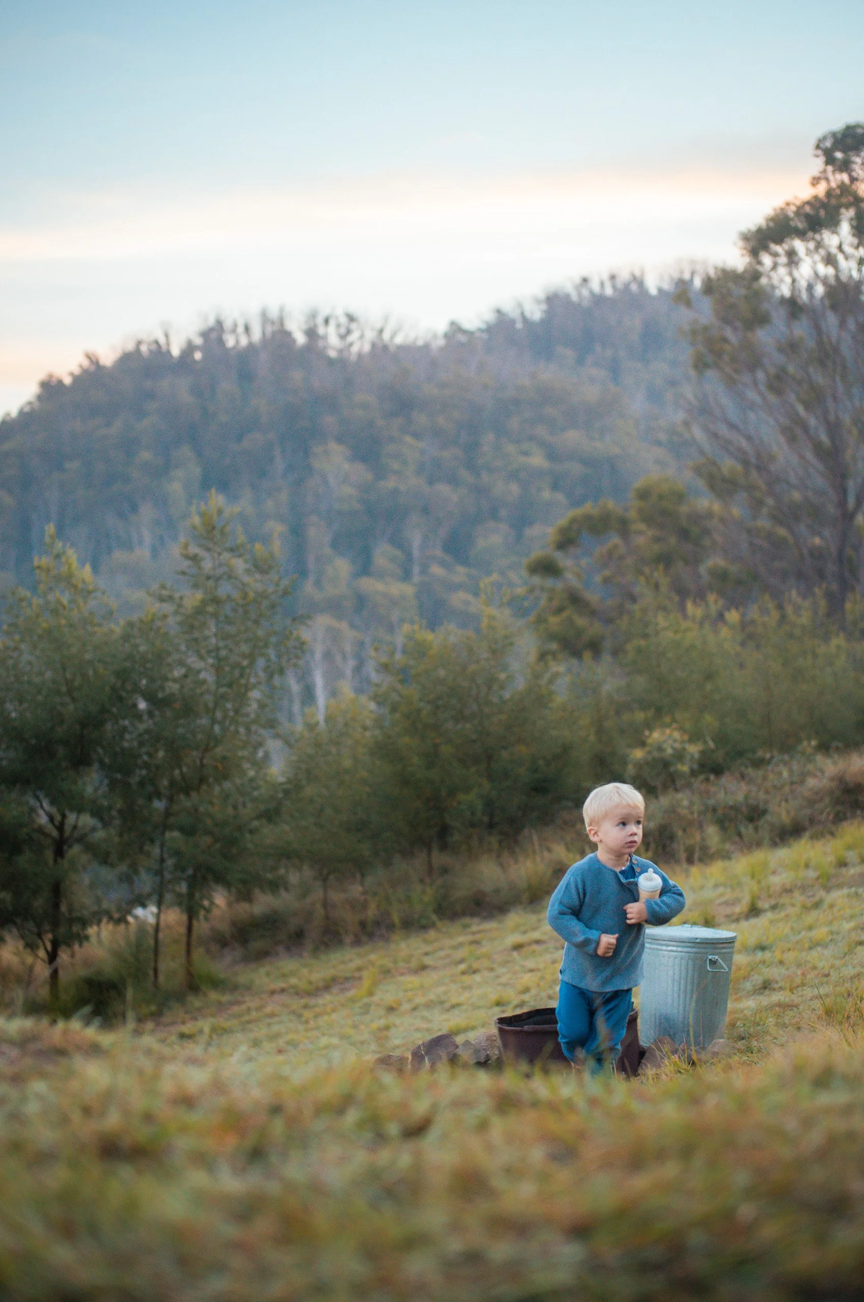 A young child with blond hair wearing a blue jacket and pants standing outdoors in a grassy field, holding a bottle. There is a metal container and a bag nearby. In the background, there are trees and hills under a partly cloudy sky.