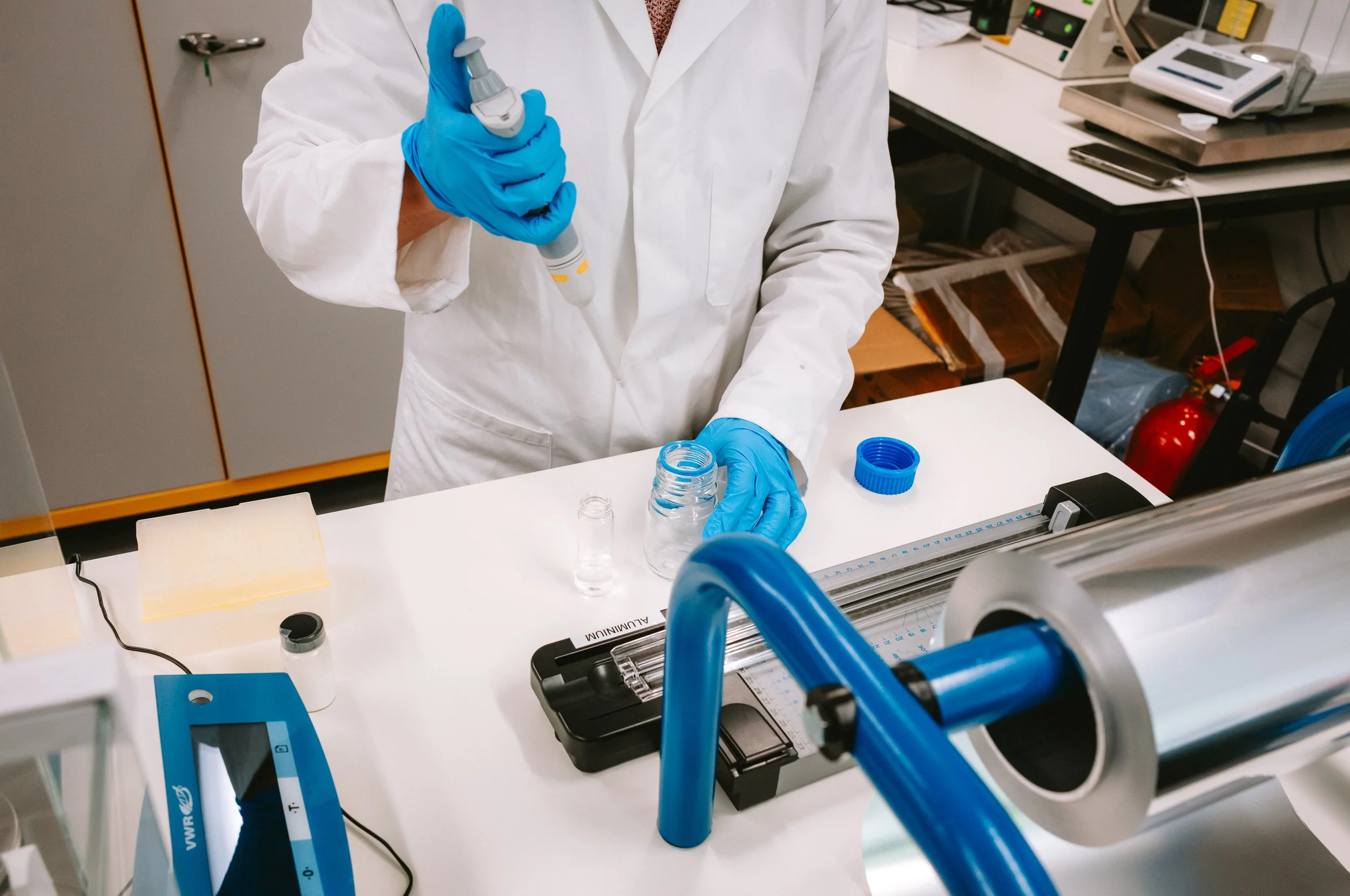 A scientist wearing a white lab coat and blue gloves using a pipette to transfer liquid into a small container in a laboratory.