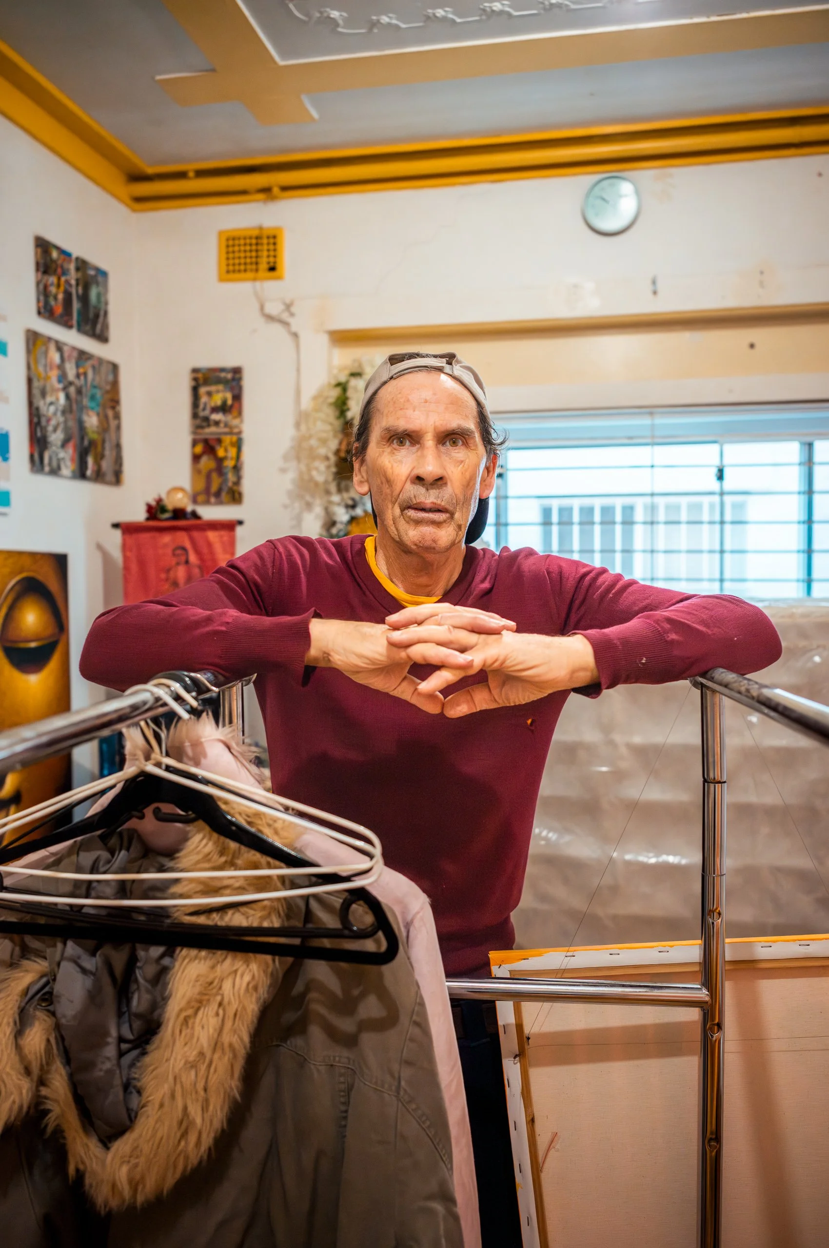 An elderly man with a distressed expression leaning on a metal rail in a room decorated with artwork and a clock, with a window in the background.