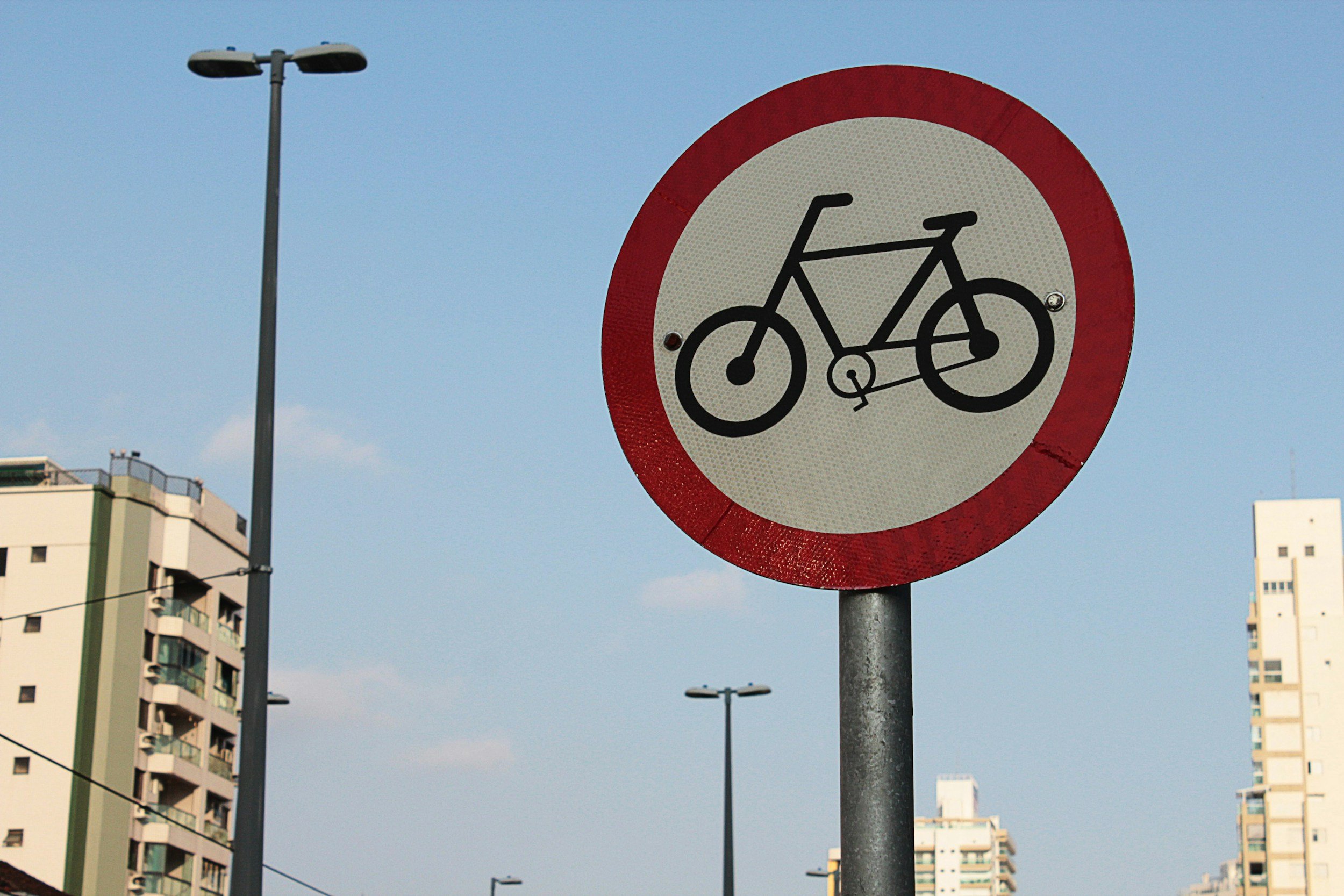 A round traffic sign indicating no bicycles allowed, with a red border and black bicycle icon on a white background. Buildings and streetlights are visible in the background under a clear blue sky.