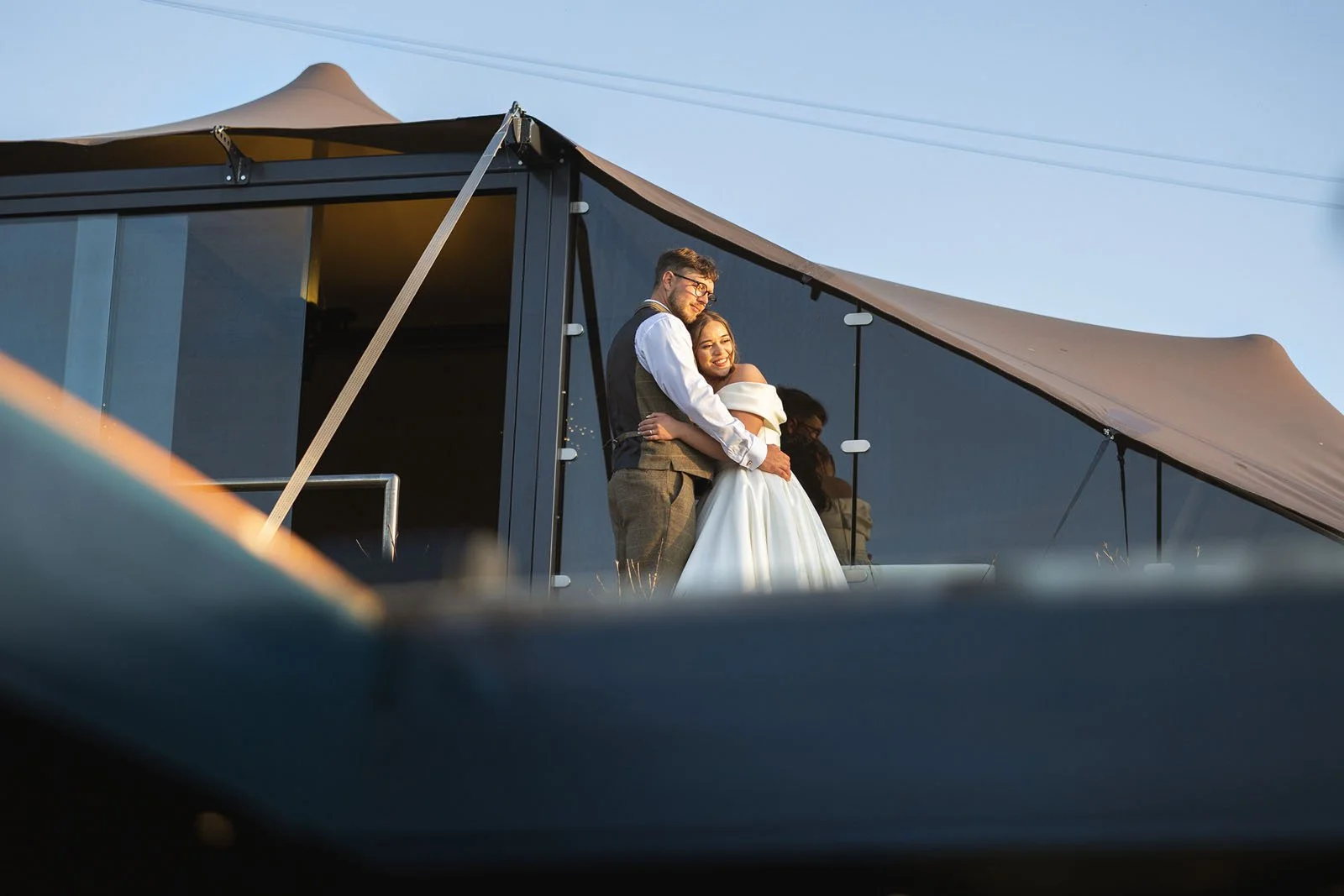 A newlywed couple hugging on a terrace or balcony during sunset, with a dark building and blue sky in the background.