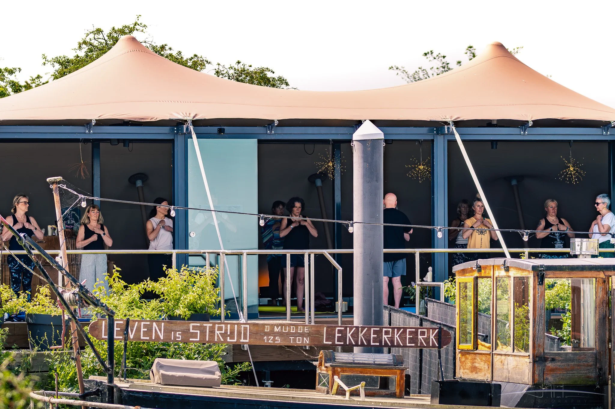 People standing inside a glass-walled building with a beige tent-like roof, surrounded by greenery and a boat in the foreground, and a sign with Danish words and weights in front.