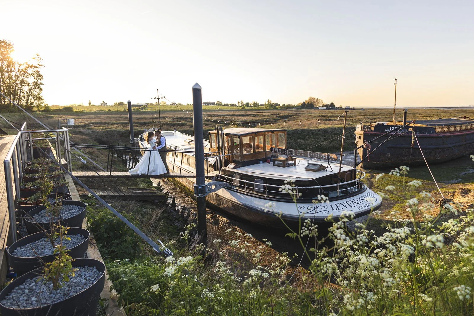 A bride and groom dancing on a dock next to a boat in a scenic outdoor setting during sunset.