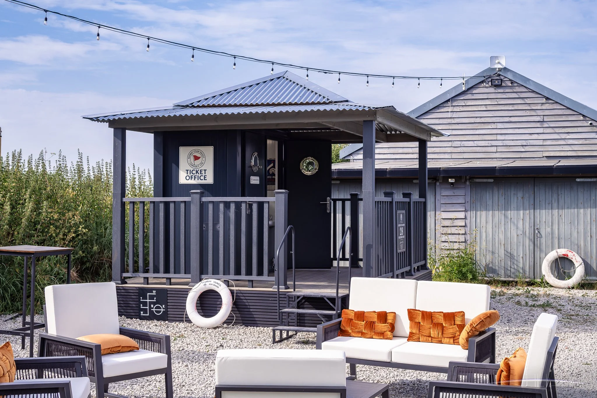 Outdoor seating area with white chairs, orange cushions, a white sofa with orange cushions, and a small black table, in front of the toilet building with a ticket office sign, under string lights, with greenery and a blue sky in the background.