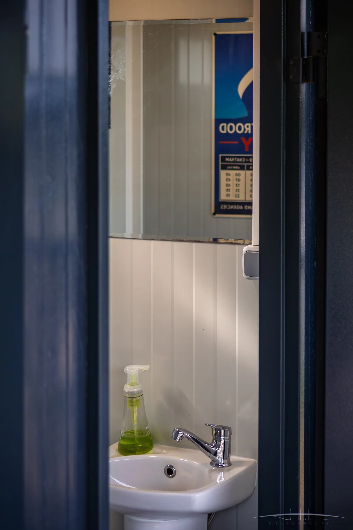 View of a bathroom sink with a soap dispenser filled with green liquid soap, a faucet, and a mirror above. The mirror reflects a sign and part of a wall.