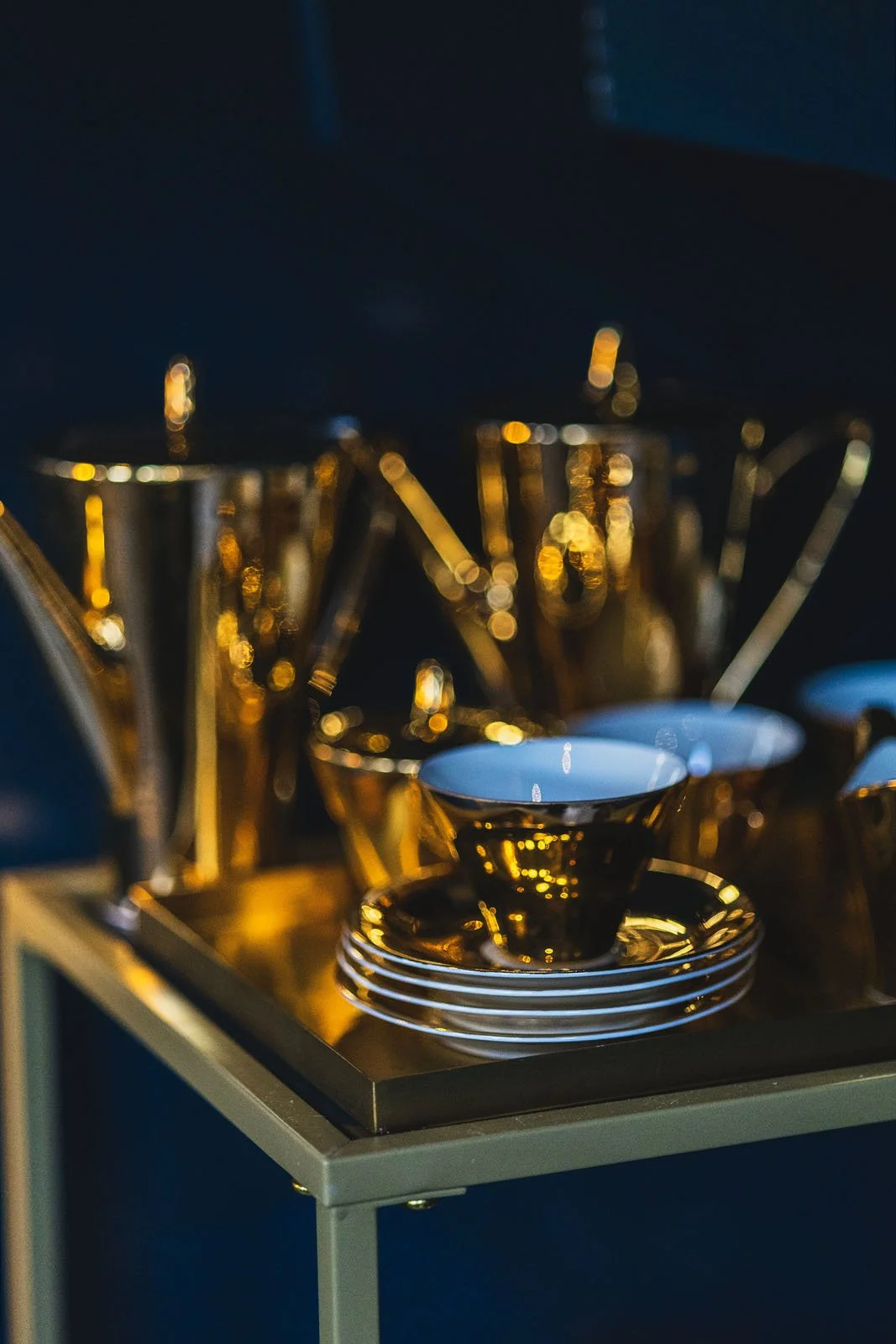 A tray with gold and white teacups, saucers, and gold utensils on a dark table with a dark background.