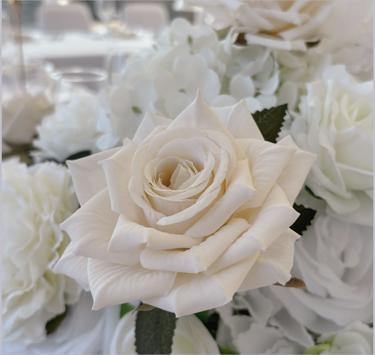 Close-up of a white artificial rose with green leaves surrounded by white flowers in the background.