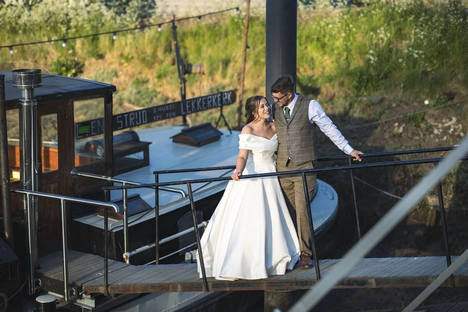 A bride and groom standing on a boat deck, smiling at each other during their wedding photoshoot in an outdoor setting with greenery in the background.