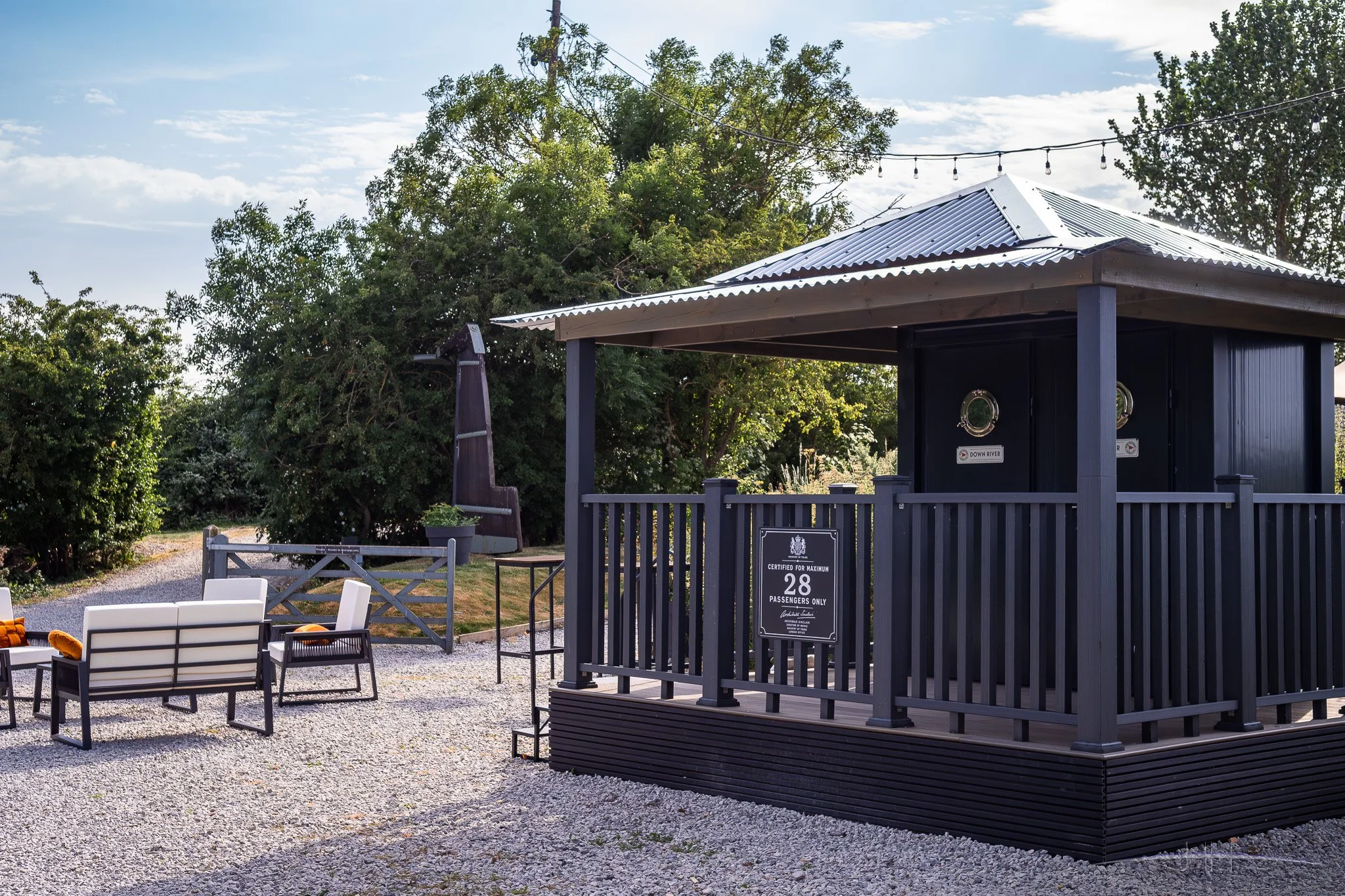 Outdoor seating area with chairs and tables, a newly created toilet building with a metal roof, surrounded by trees and string lights, under a partly cloudy sky.