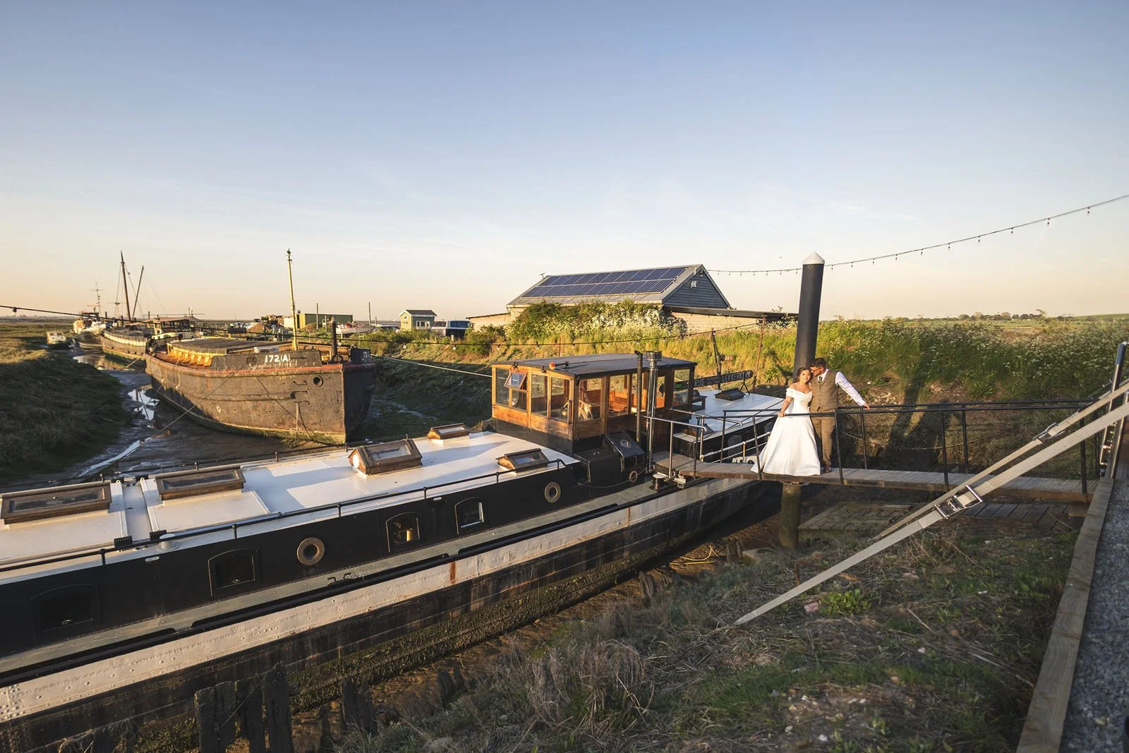 Wedding couple standing on a boat docked along a canal with boats and a building in the background during sunset.