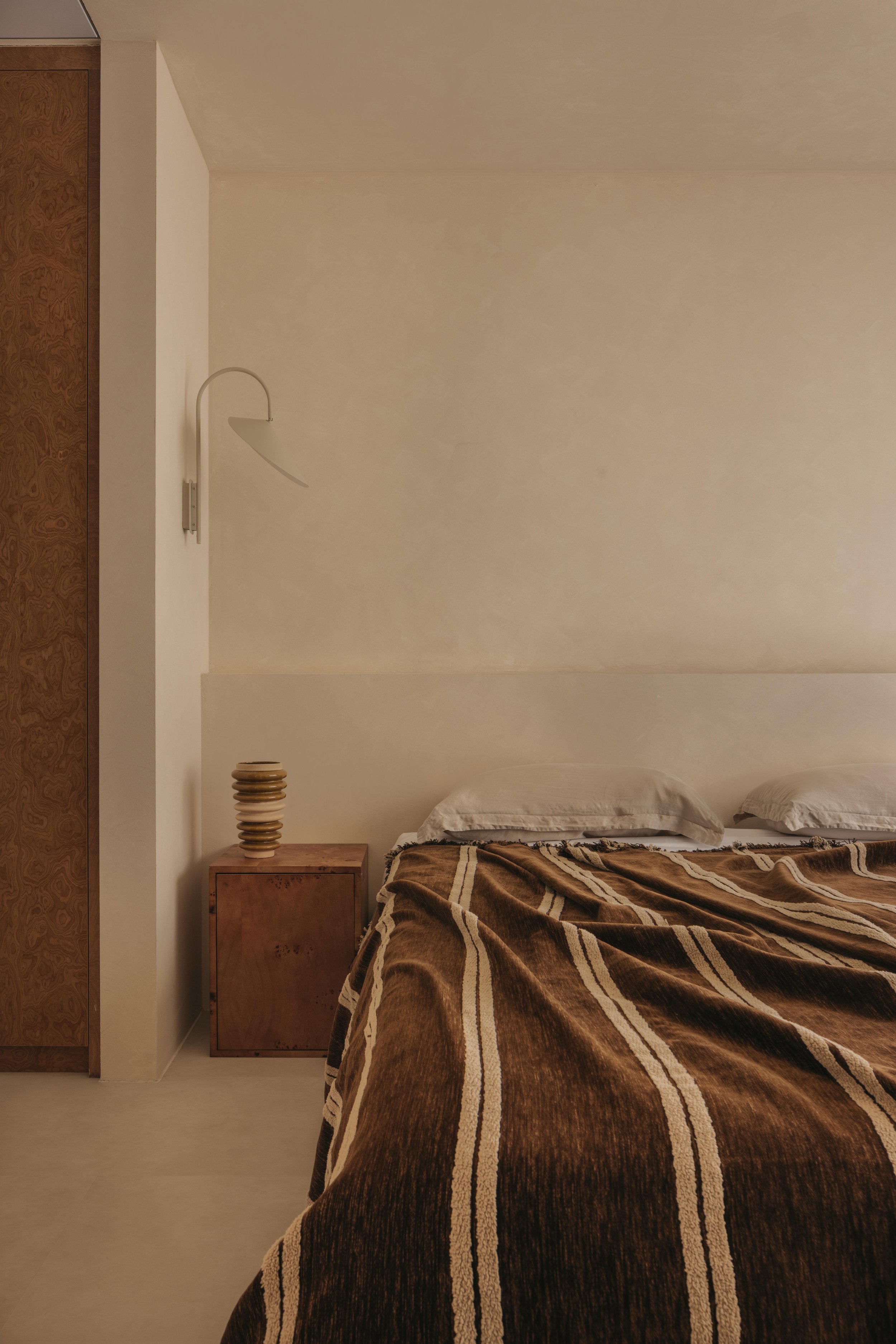 A minimalist bedroom with a wooden nightstand, a decorative wooden lamp, and a bed covered with a brown striped blanket. The wall is plain, with a reading lamp mounted beside the bed.