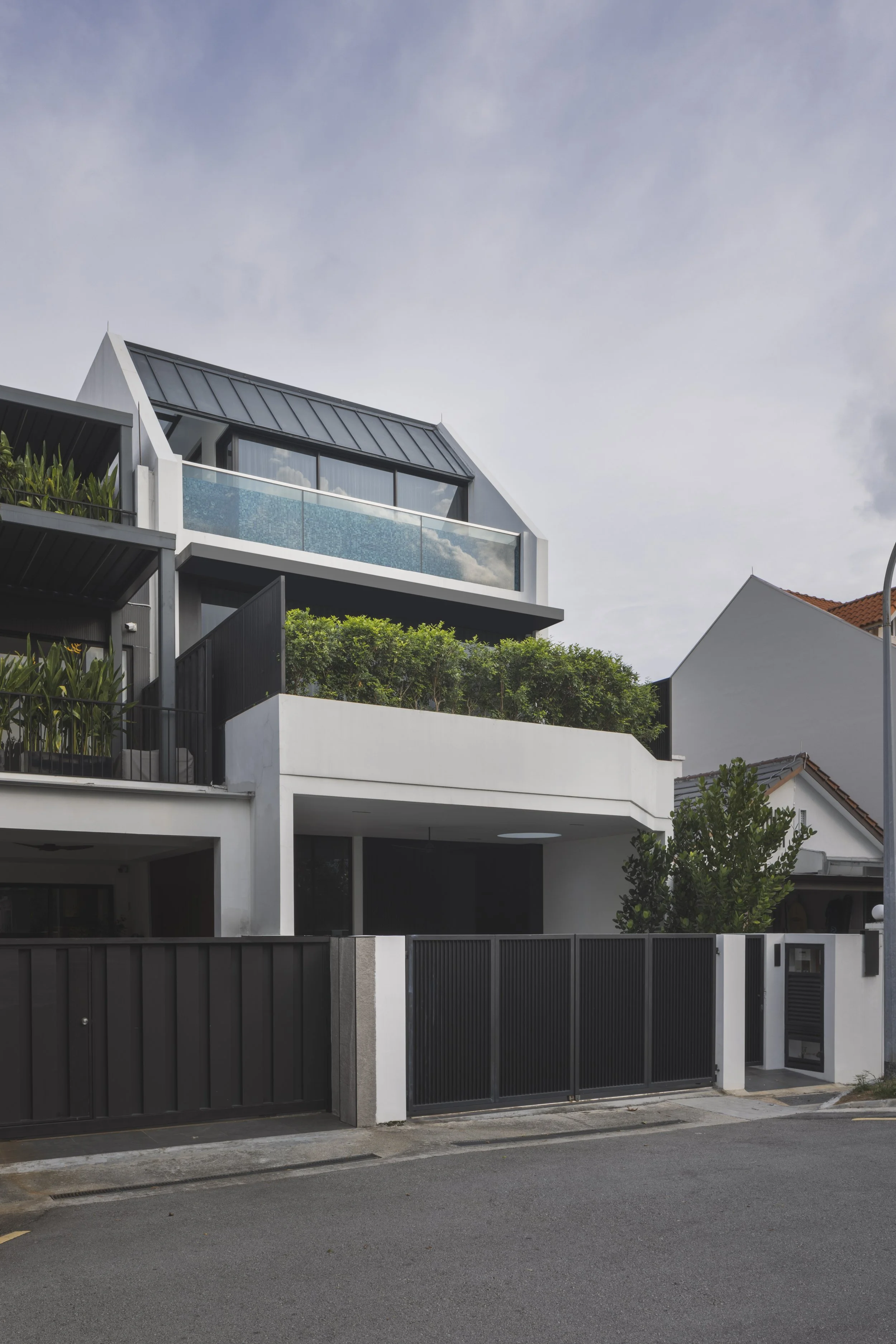 Modern white residential building with black gates, balcony with glass railing, some greenery, and neighboring white houses, under a cloudy sky.