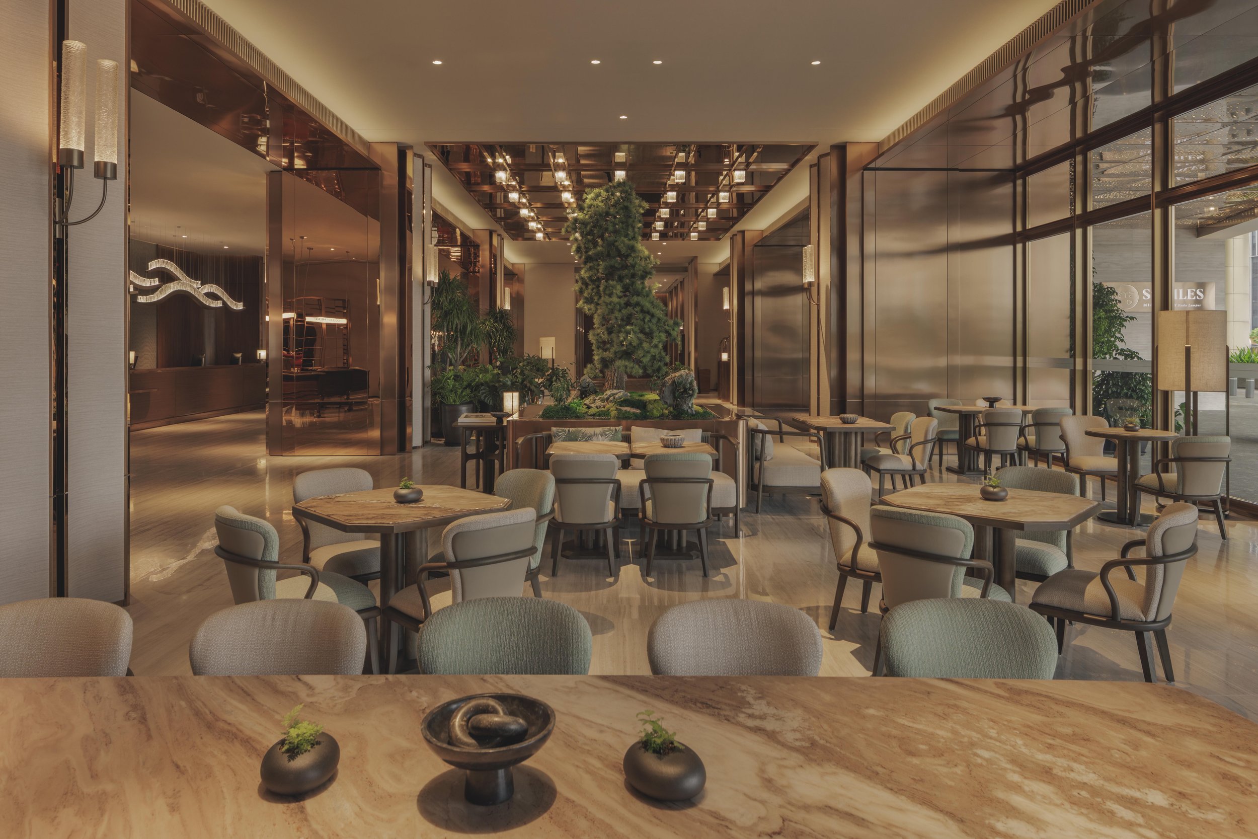 Elegant hotel lobby with a large indoor tree, modern lighting, and seating area with round tables and cushioned chairs.
