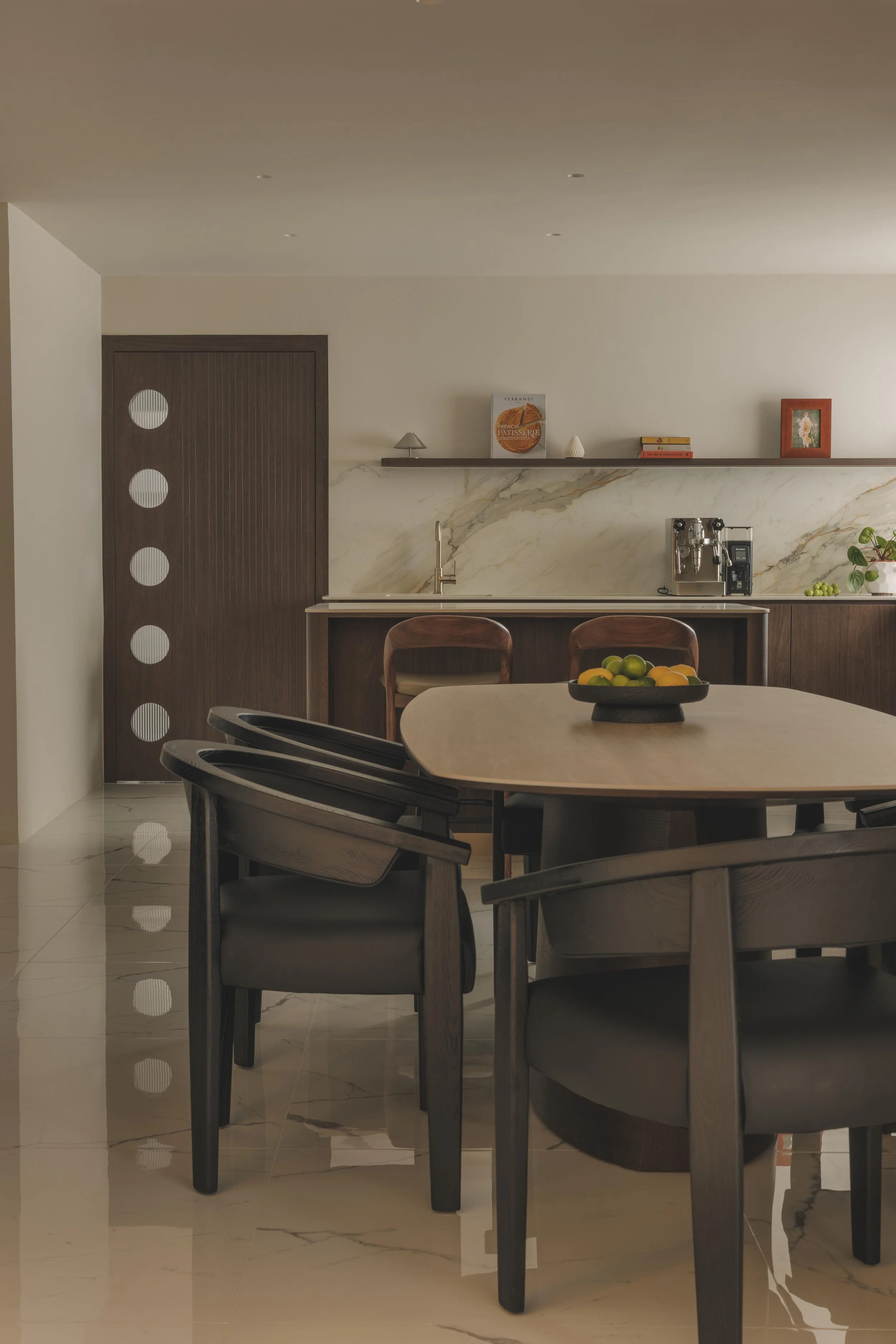 Modern kitchen with dark wood chairs around a light wooden table, a backsplash with marble pattern, a coffee machine, fruit bowl, and decorative books and artwork on a shelf.