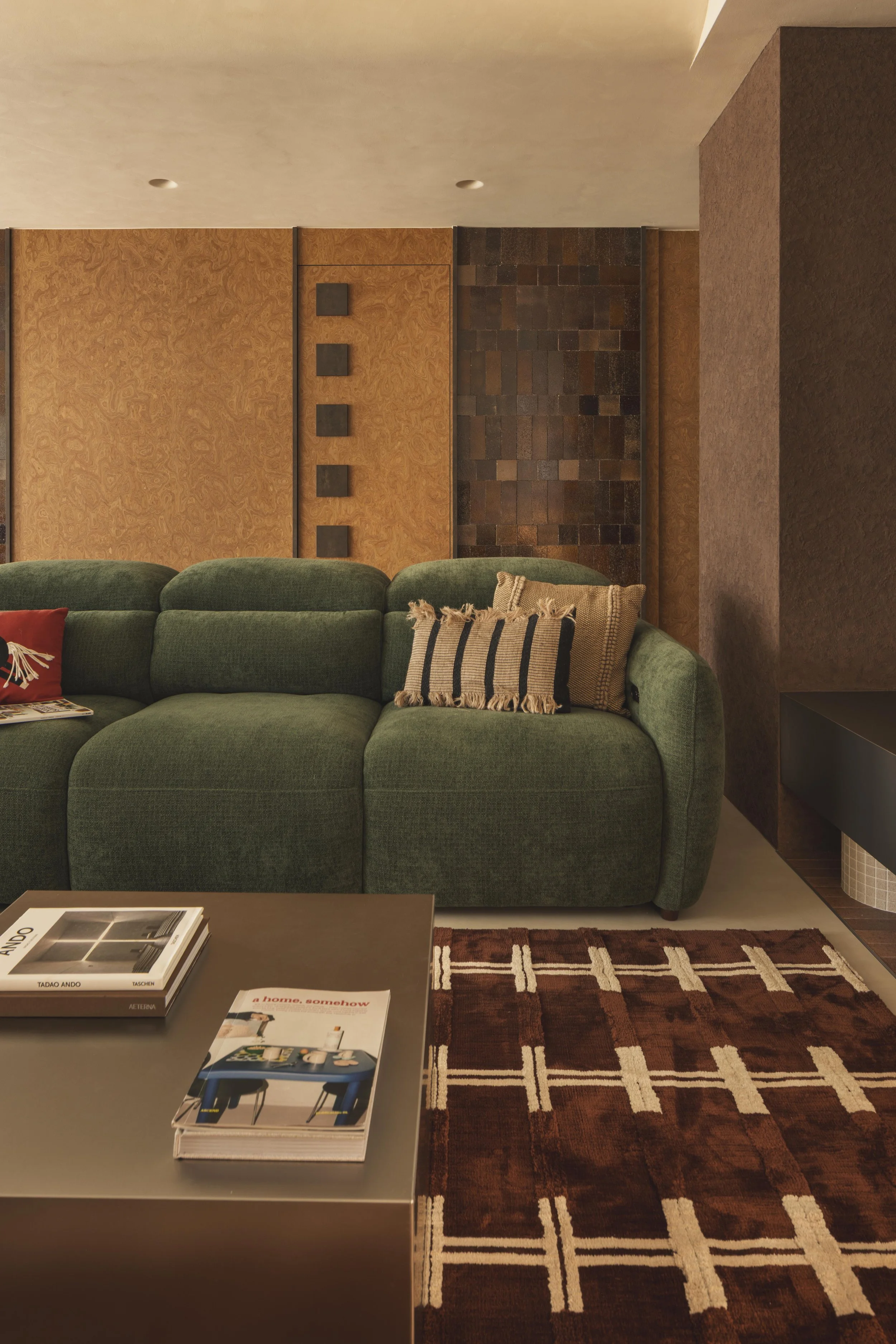 Living room with a green upholstered sofa, a brown and white geometric patterned rug, a dark gray coffee table with books on it, and a wall with wood paneling and textured brown bricks. There are also some throw pillows on the sofa and a magazine on the table.