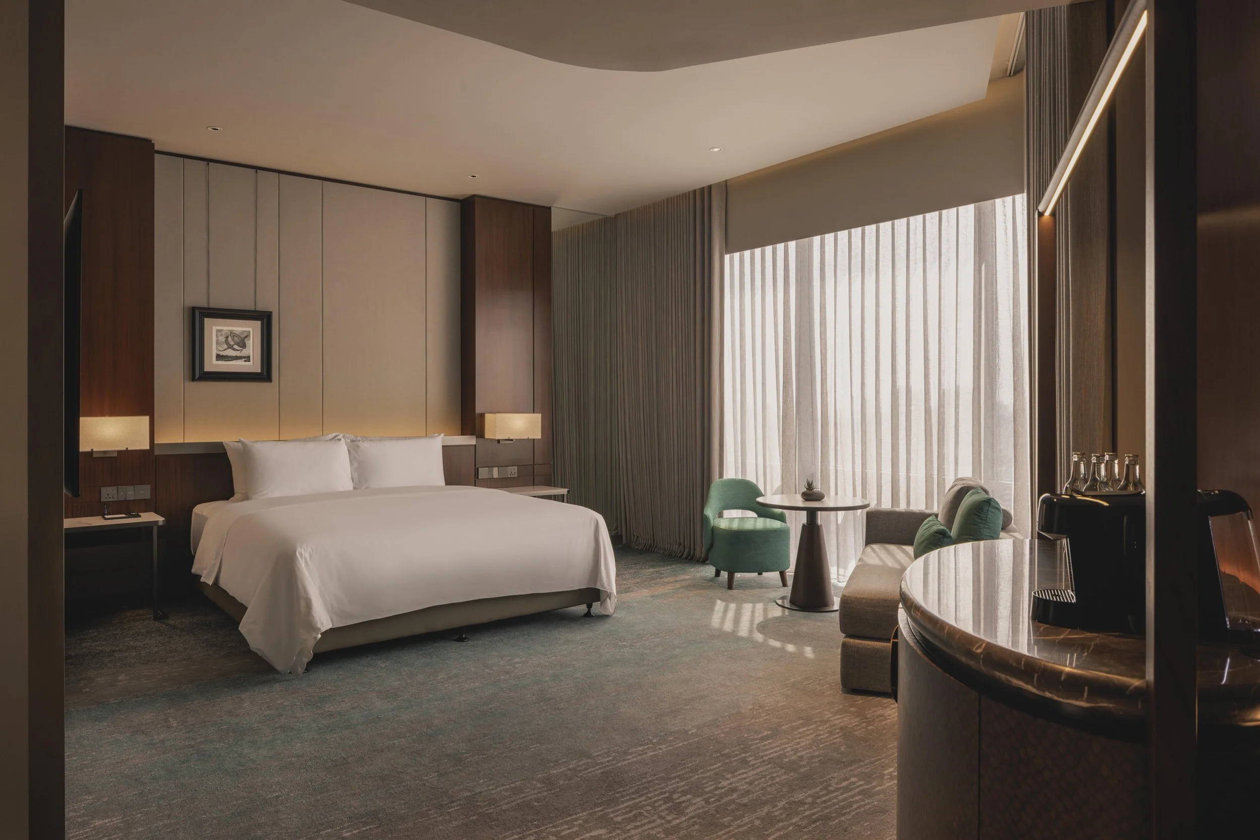 Interior of a modern hotel room featuring a bed with white bedding, a small round table with green chairs, a framed picture on the wall, beige curtains covering large windows, and warm lighting.