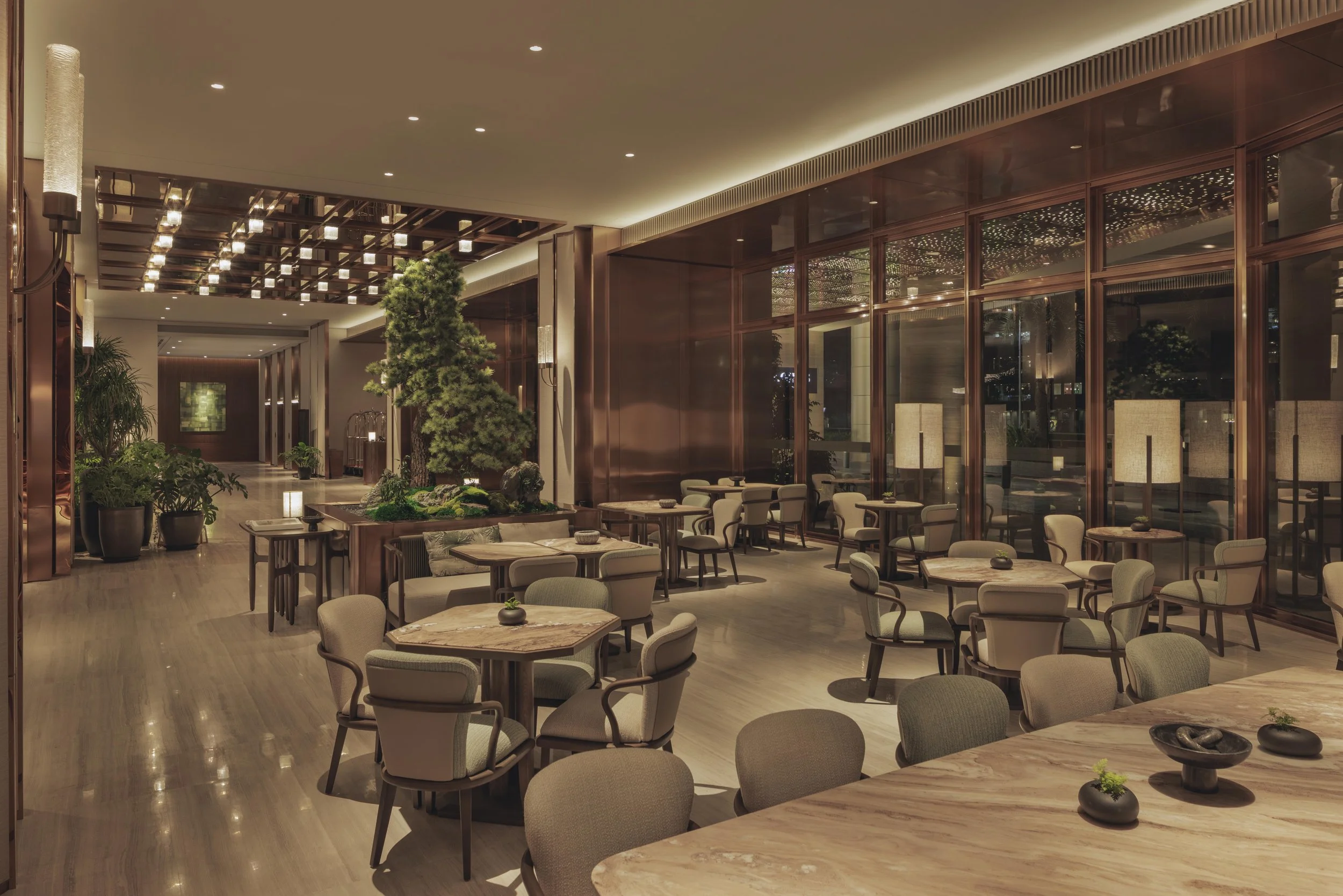 Elegant hotel lobby with warm lighting, potted plants, and seating area with beige chairs and marble tables.