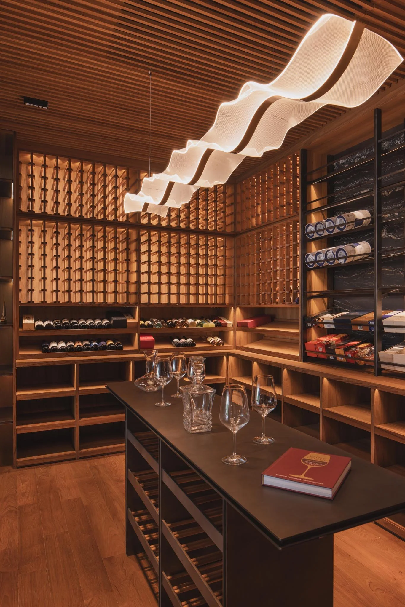 Modern wine cellar with wooden wine racks, a black table with wine glasses, and a contemporary wavy chandelier ceiling light.