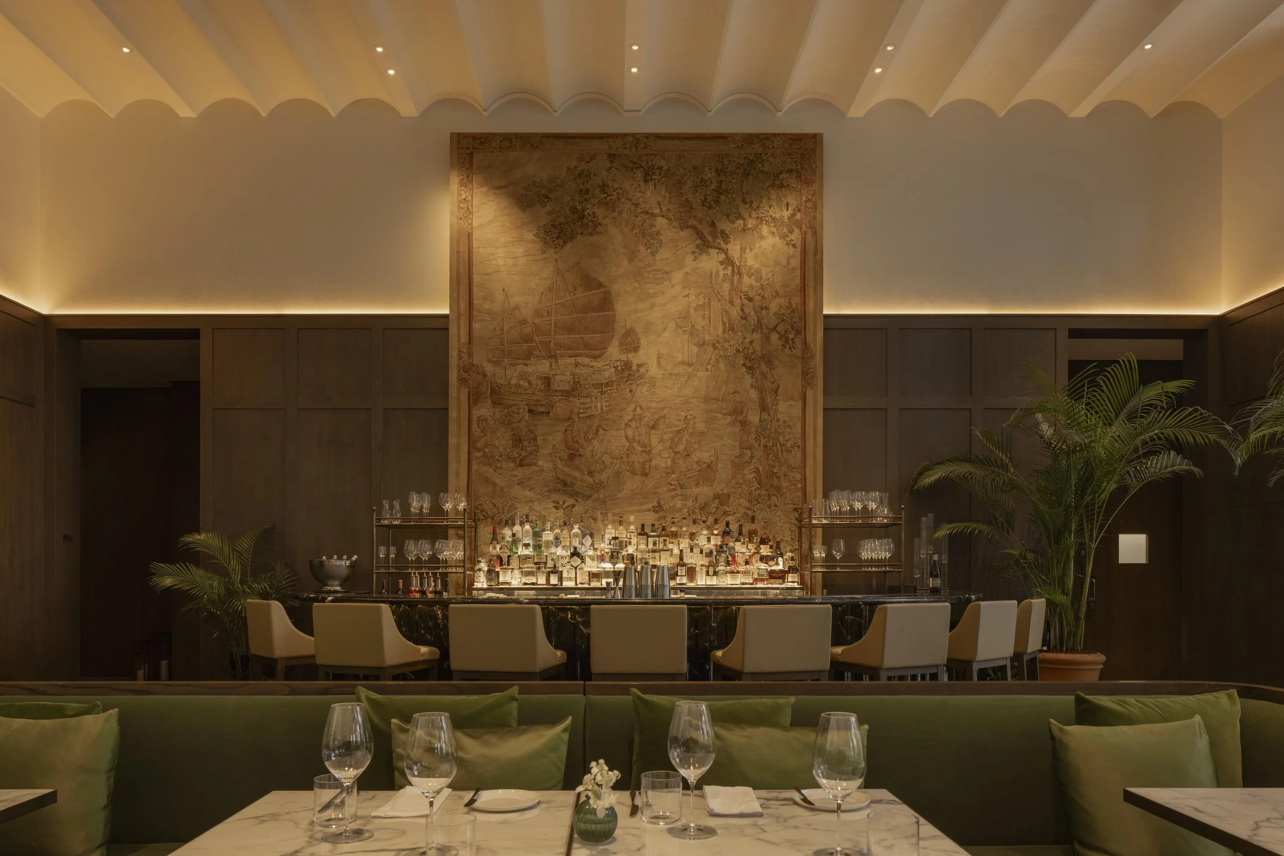 An elegant bar area with a large painted mural of a ships, surrounded by warm lighting, with a marble-topped bar counter, and set tables in the foreground.