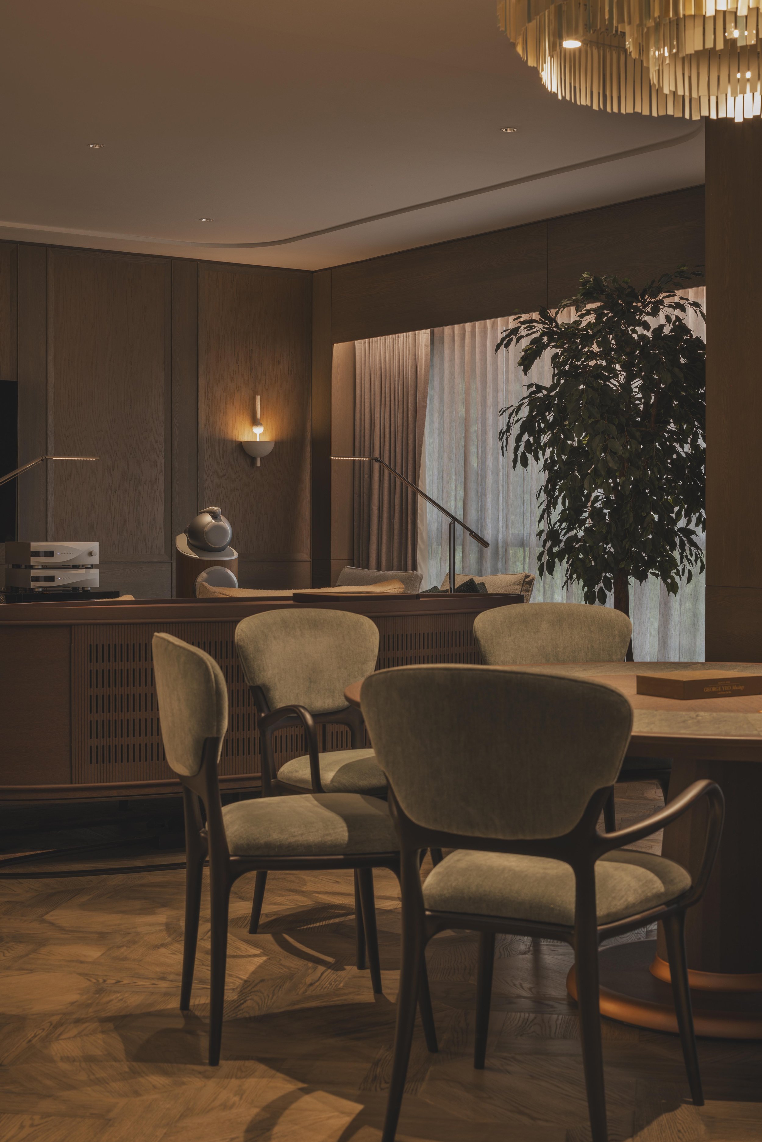 Interior of a modern living space with a round wooden table, upholstered chairs, a large potted plant, a wooden wall with built-in lighting, and a window with curtains.