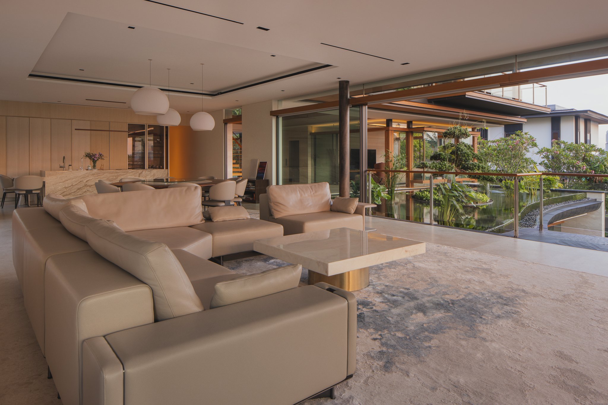 Modern open-concept living room with beige sofas, marble coffee table, and large windows overlooking a garden with lush greenery and water feature.