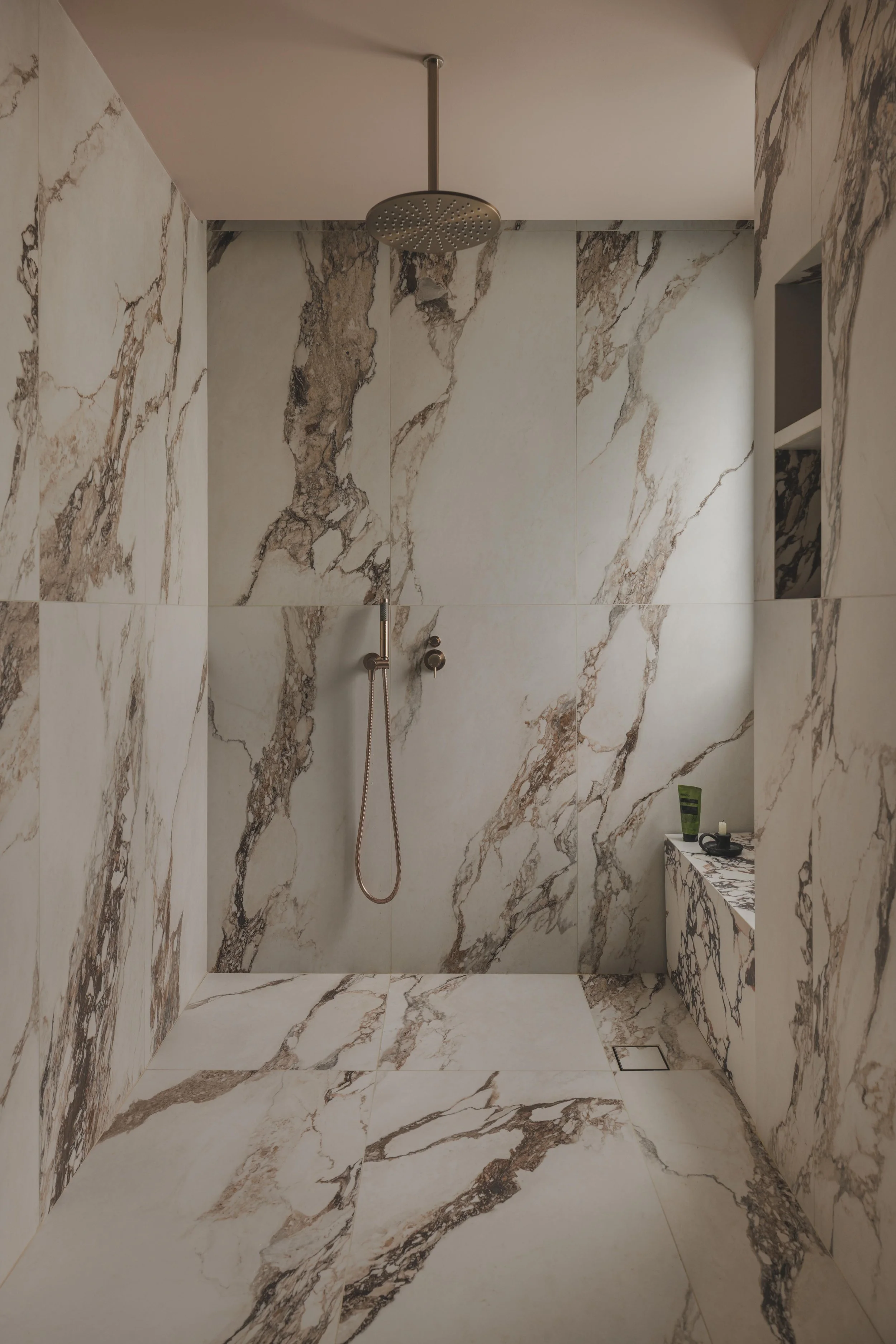 A modern bathroom with walls, floor, and ceiling covered in beige marble with brown veining, featuring a rainfall showerhead, a handheld shower, and a small built-in shelf with toiletries.