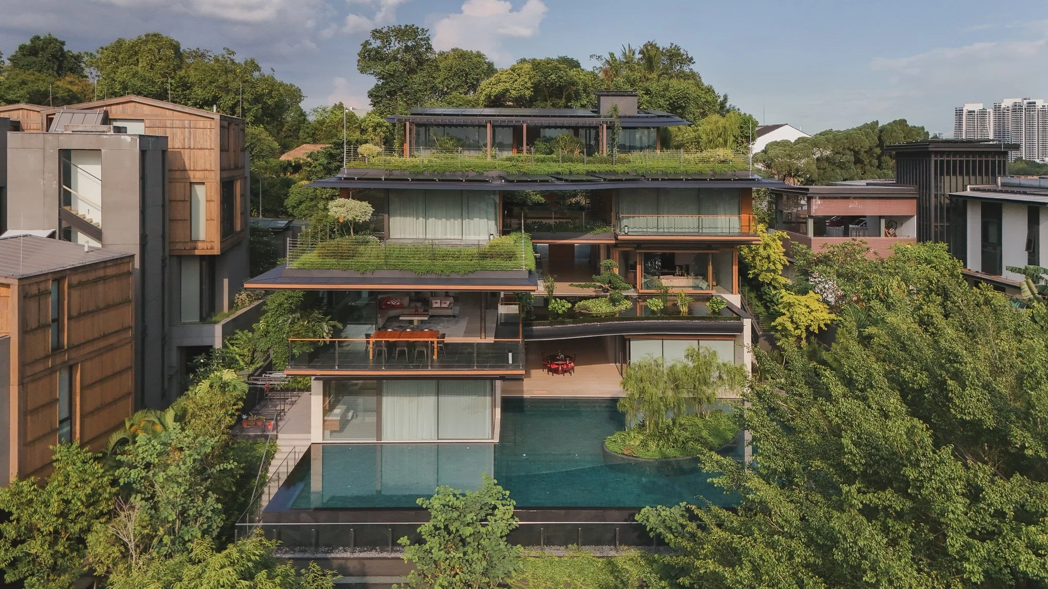 A modern multi-story house surrounded by lush greenery, with terraces, large glass windows, and a swimming pool.