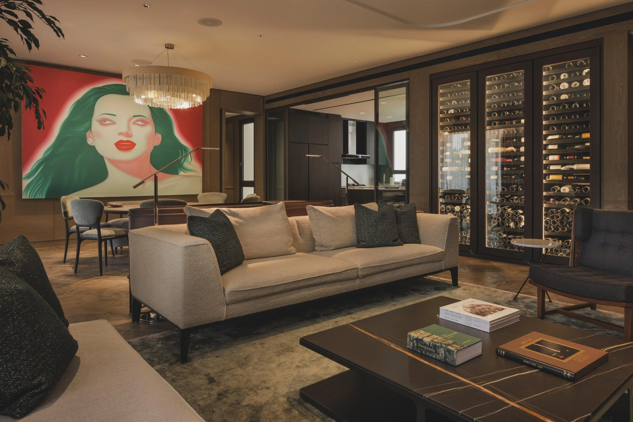 Living room with beige sofa, black coffee table with books, and a large wall art of a woman with green hair and red lips on a red background. There is a drum chandelier hanging from the ceiling, and wine storage near a glass wall.