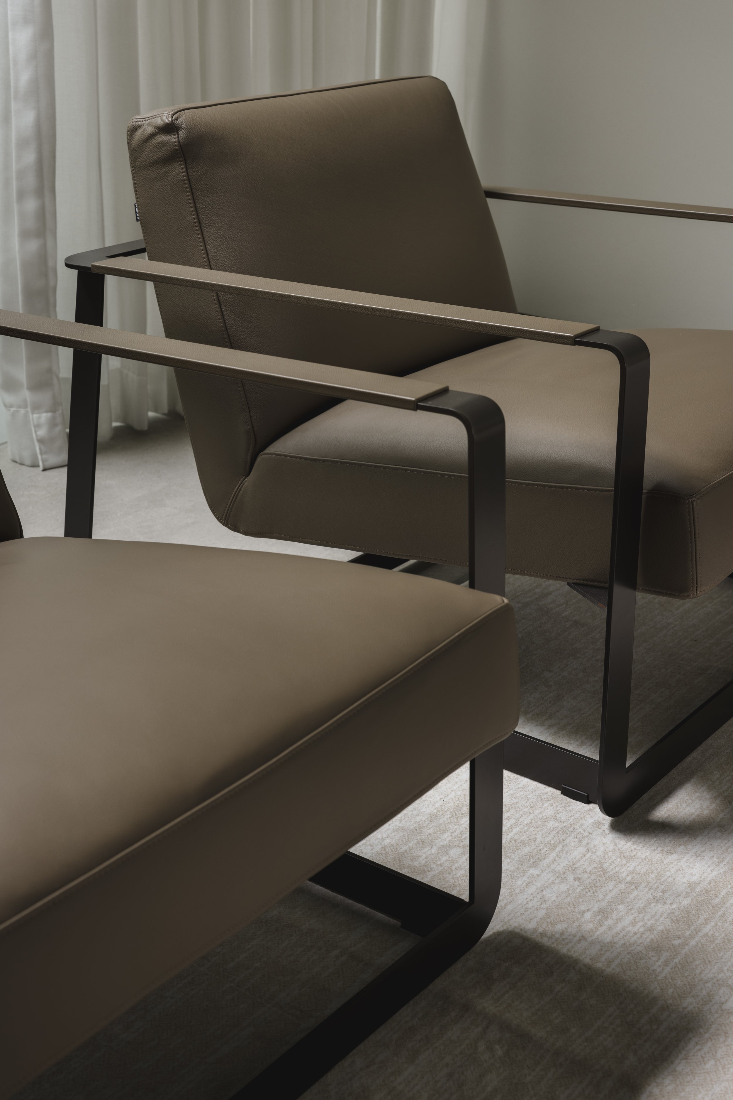 Close-up of a modern armchair with dark metal frame and beige cushioned seat and backrest, placed near a window with light curtains.