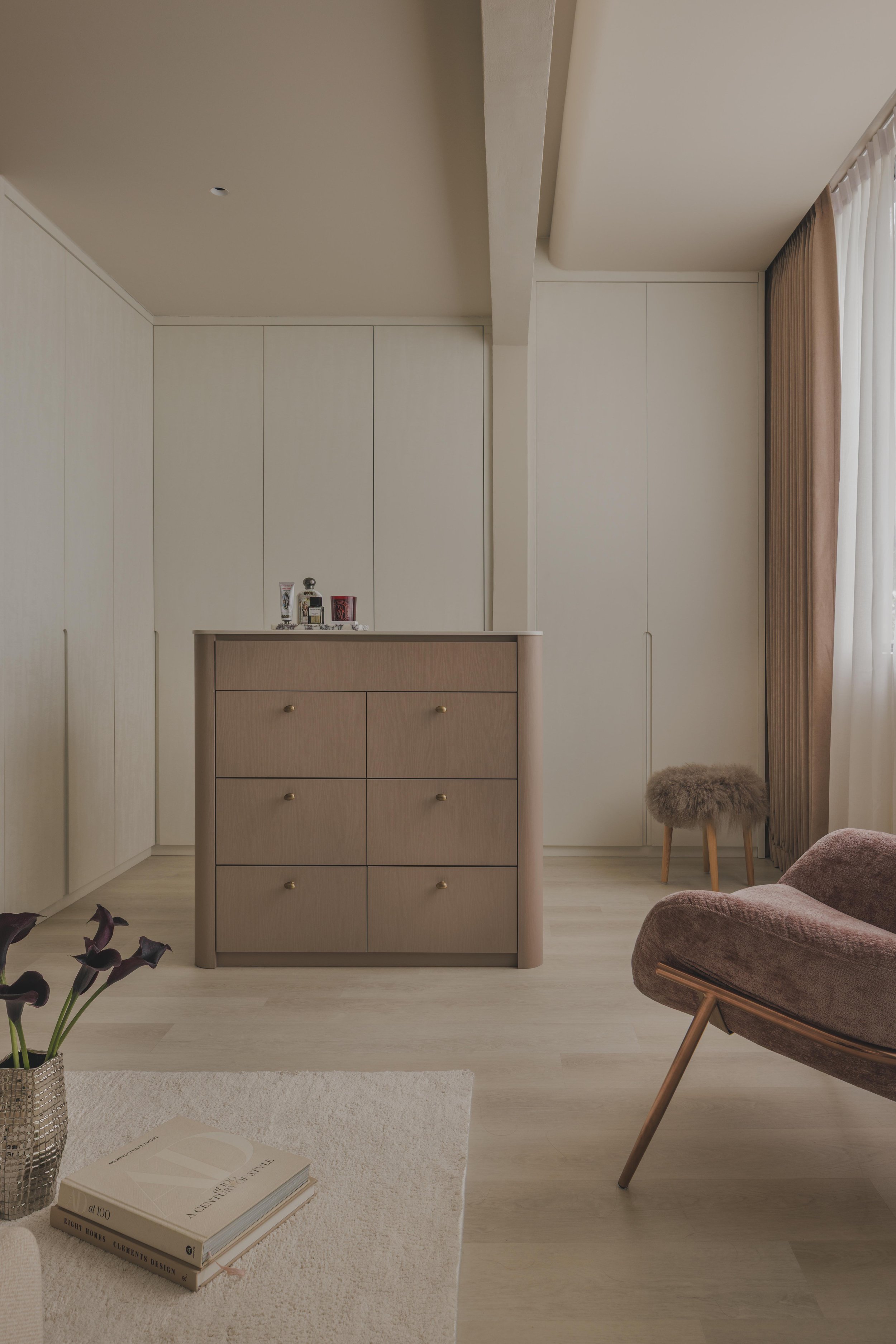 A cozy living room corner with a beige dresser, a soft armchair, a small stool, and a vase with dark purple flowers. Books are on a white rug on the light wood floor, and a window with pink and white curtains is on the right side.