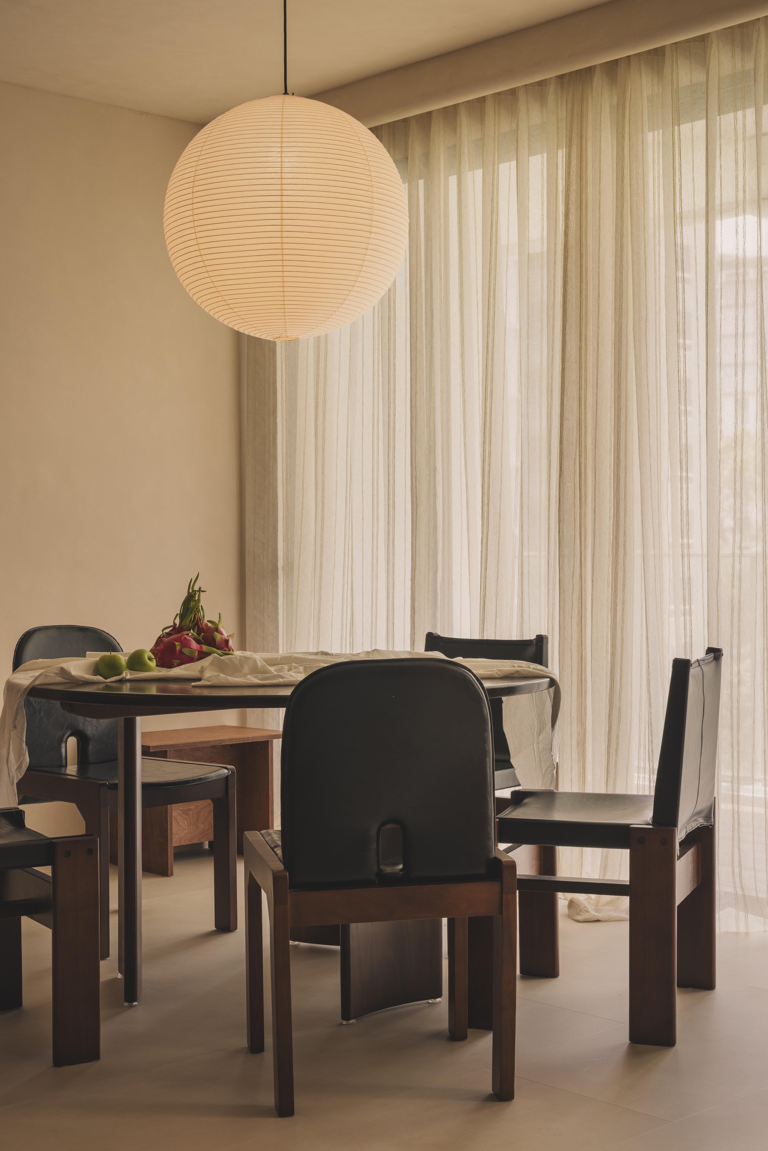 A dining room with a round wooden table, surrounded by black and wooden chairs. The table has a white tablecloth and a floral centerpiece with pink and red flowers and green apples. The room has beige curtains covering large windows, a paper lantern hanging from the ceiling, and light-colored walls.