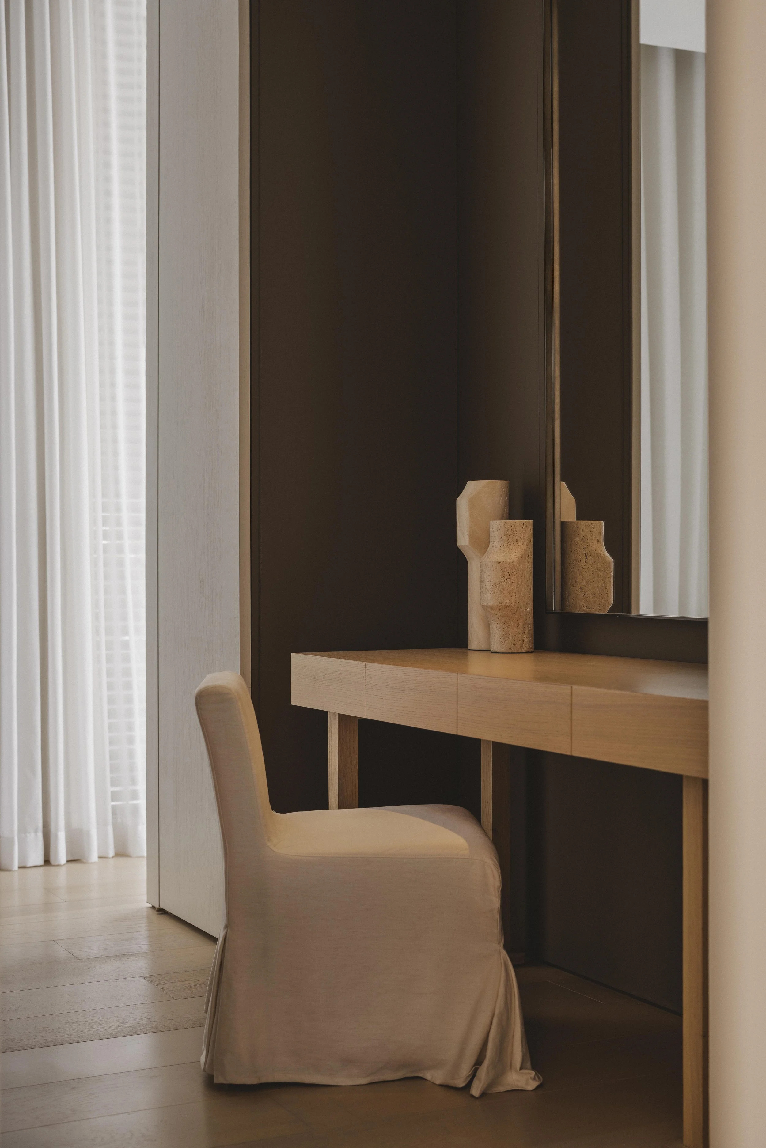 Corner of a room with a wooden desk with decorative vases, a beige upholstered chair, dark wood cabinet, cream curtains, and wooden flooring