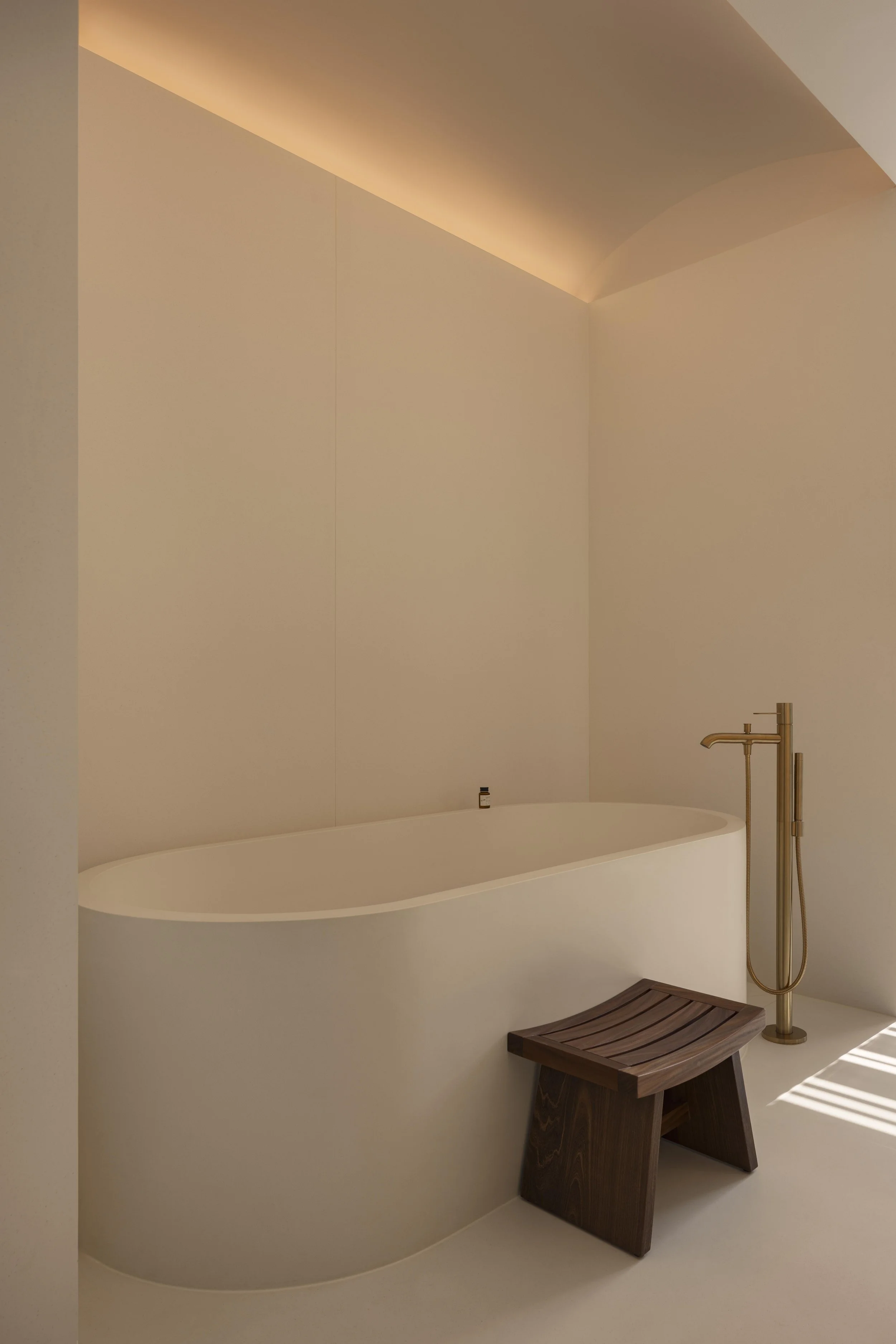 A modern bathtub with a sleek design, a brass floor-mounted shower faucet, a small bottle of bath product on the tub's edge, a wooden bench, and soft warm lighting