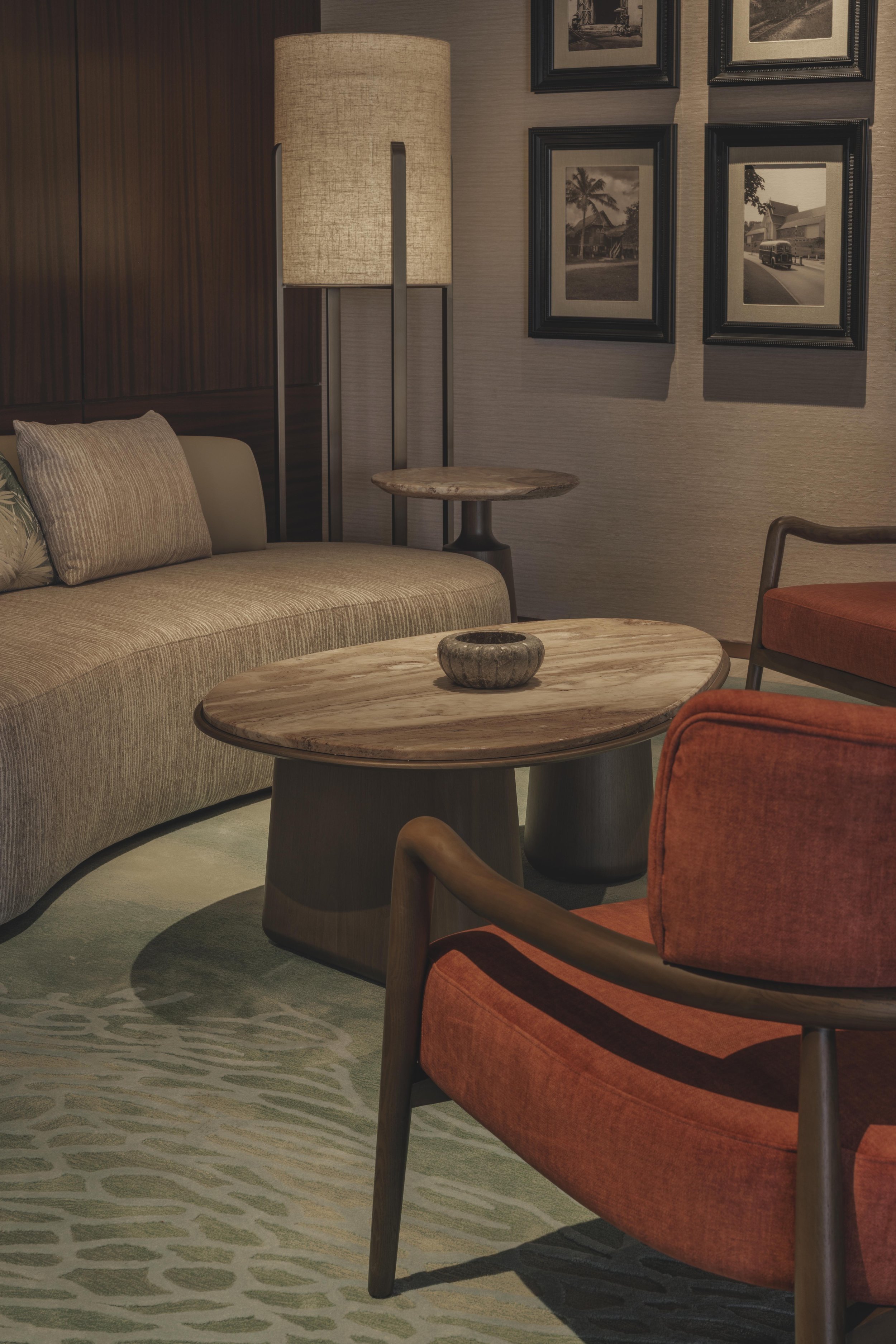A cozy hotel lounge area with a beige sofa, orange armchair, circular wooden coffee table, small side table, and decorative framed black-and-white photographs on the wall. A tall lamp with a beige shade provides warm lighting, and a patterned rug covers the floor.