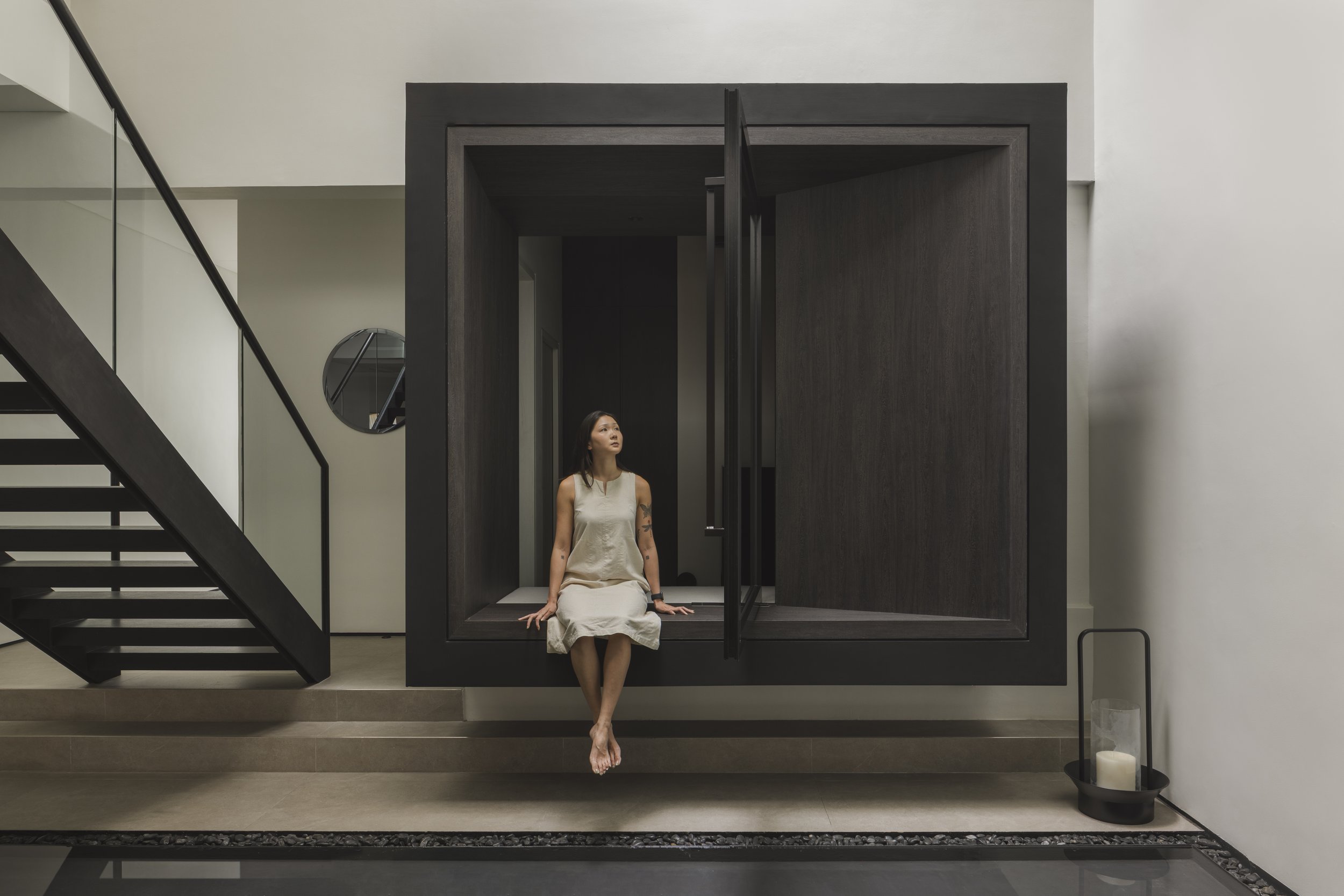 A woman in a beige dress sits cross-legged in a modern, minimalist interior space within a dark wooden frame structure. There is a staircase on the left side of the image, a round mirror on the back wall, and a black tray with a white candle on the right side.