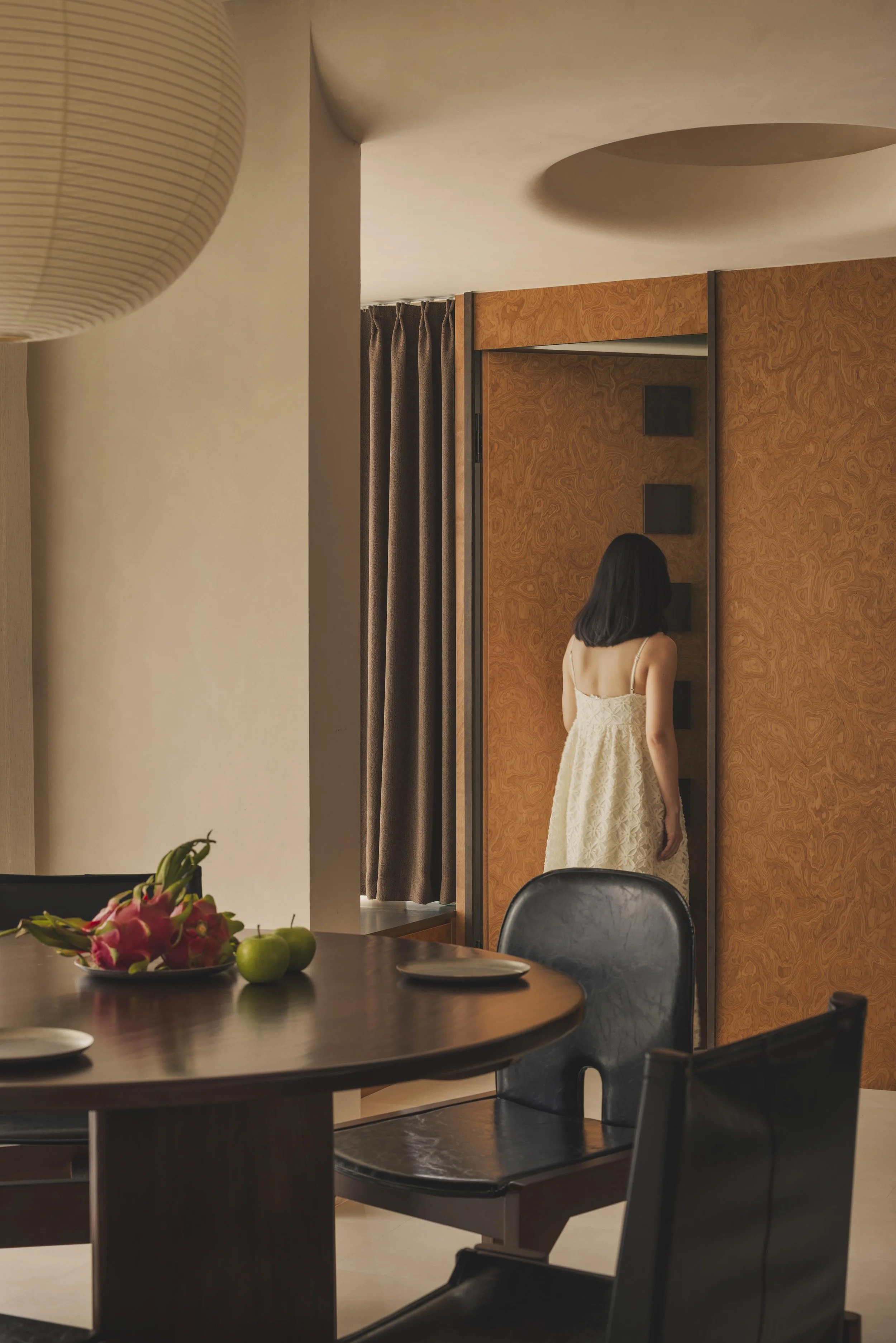 A woman in a cream-colored dress is standing with her back to the camera, inside a room with wooden walls and dark curtains. There is a dining table in the foreground with a bowl of pink lilies and green apples, and black chairs surrounding it.