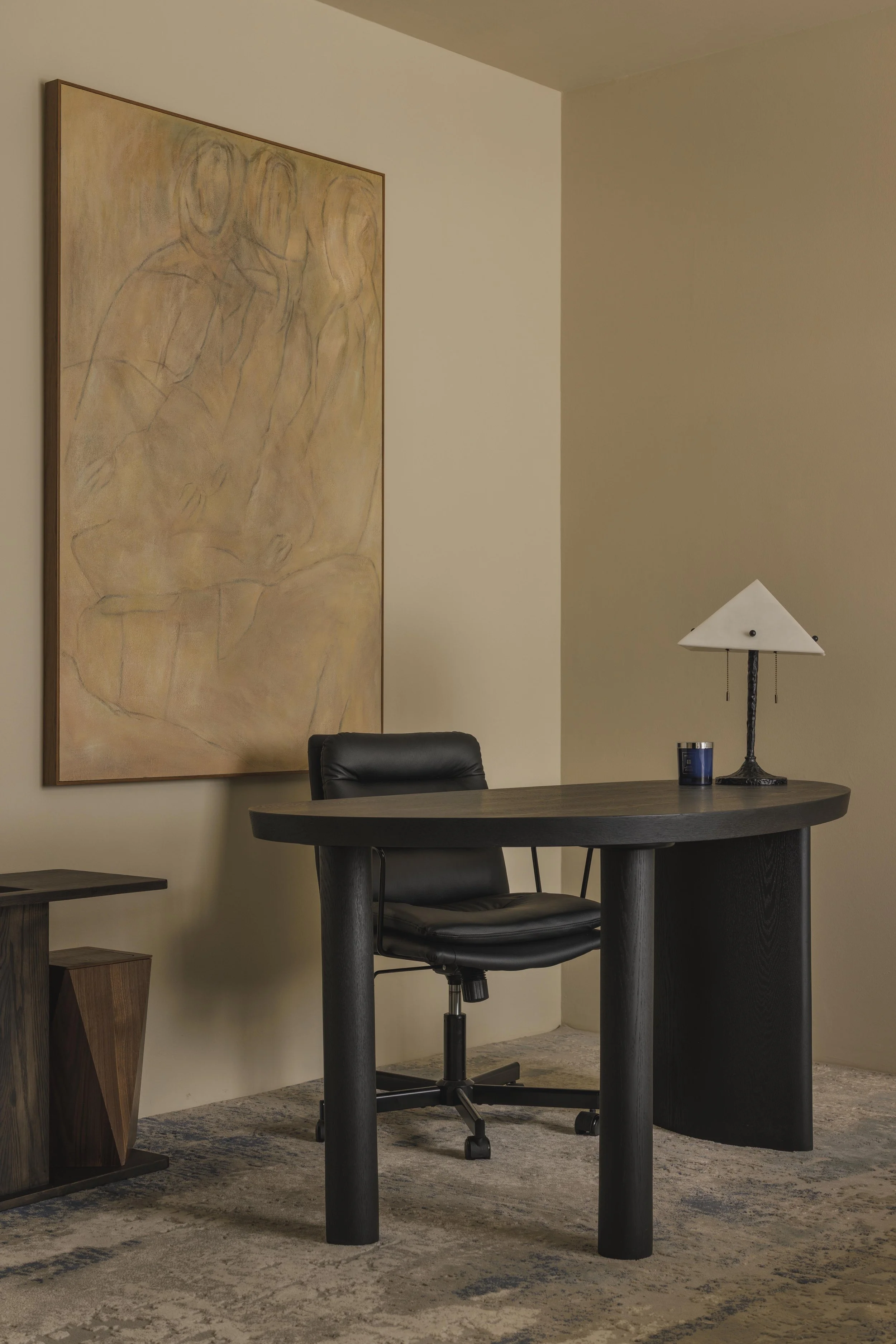 A modern office corner with a round dark wood table, black ergonomic chair, a lamp with a white shade on a black stand, a small blue cup, and abstract art on the beige wall.