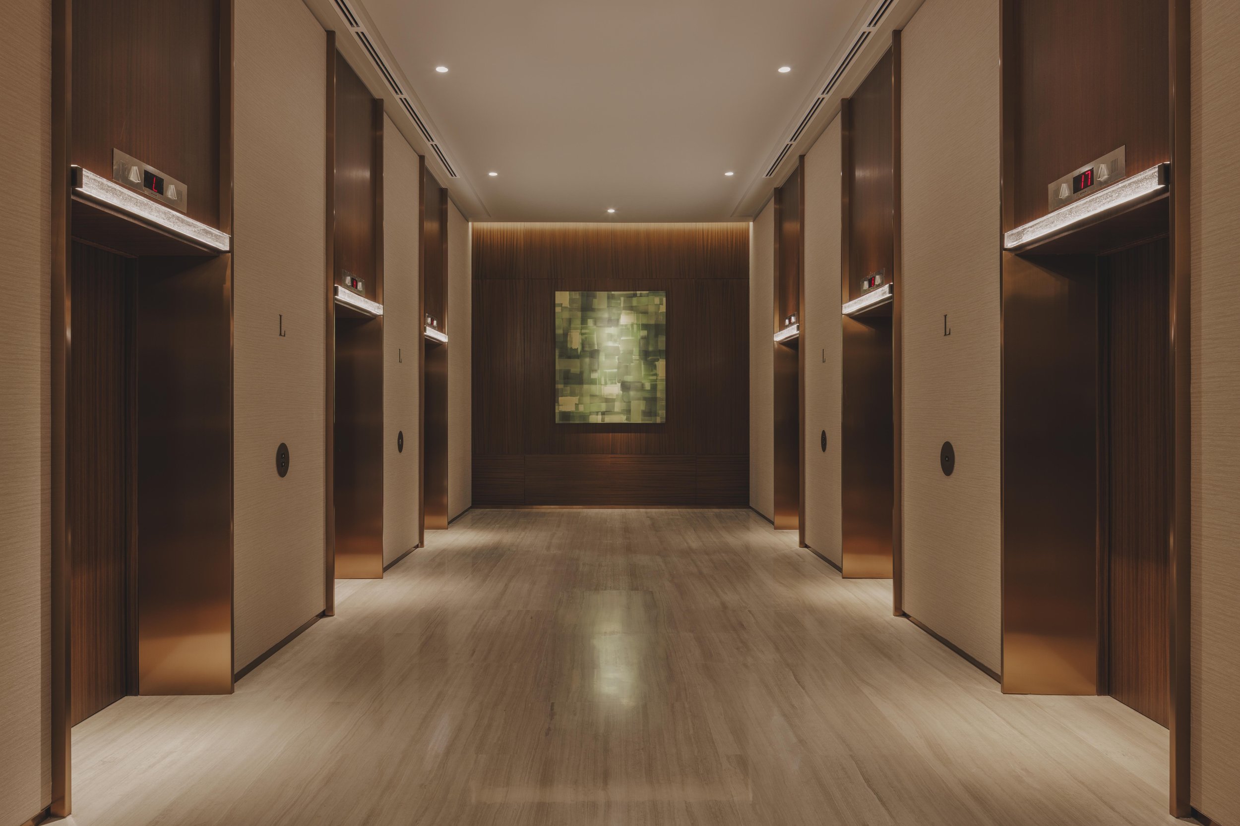 Elevator lobby with four elevators, wooden panel doors, dim lighting, beige walls, and a framed abstract art piece on the back wall.