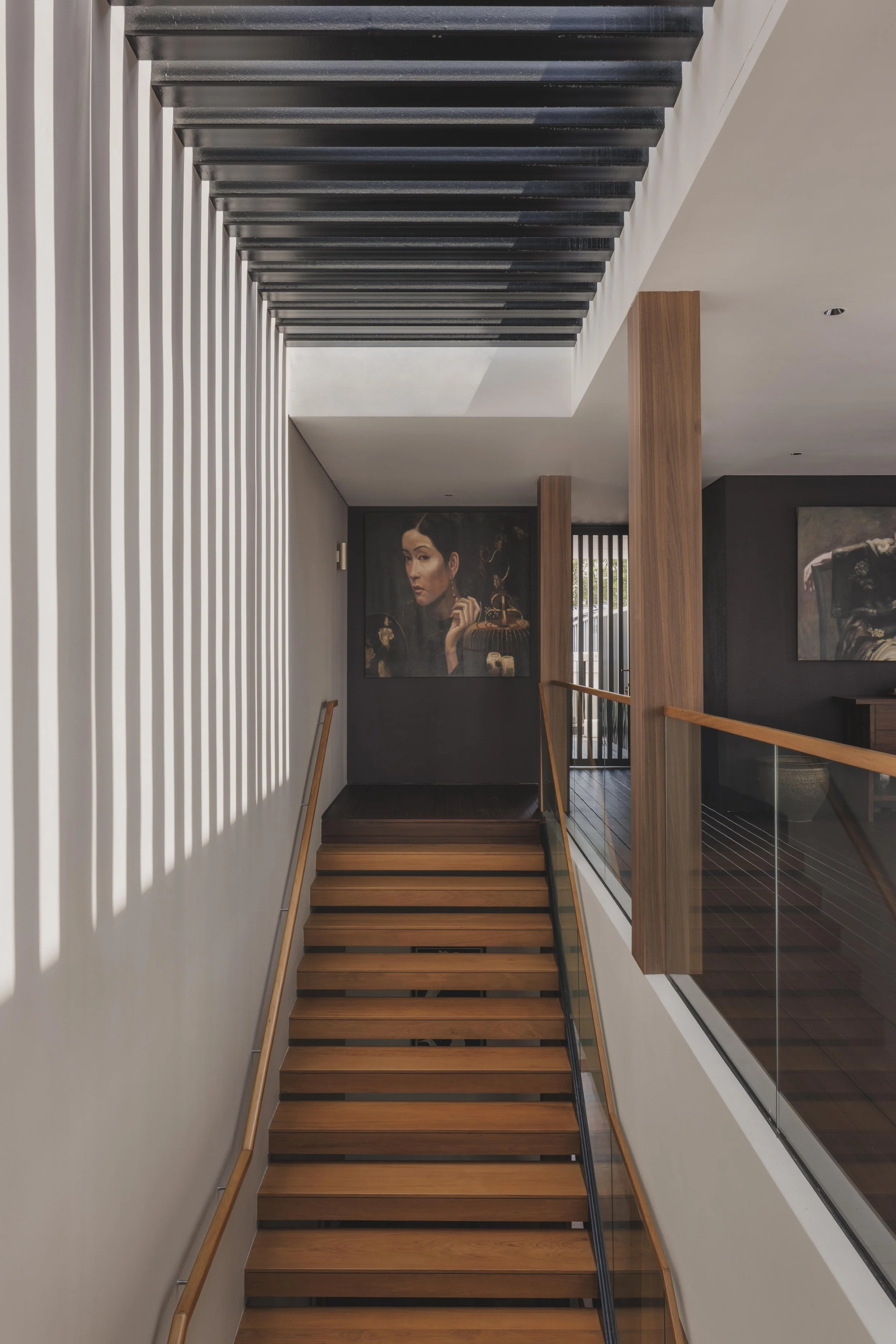 Interior view of a modern staircase with wooden steps, glass railing, white walls, and paintings on the wall in a contemporary home or art gallery.